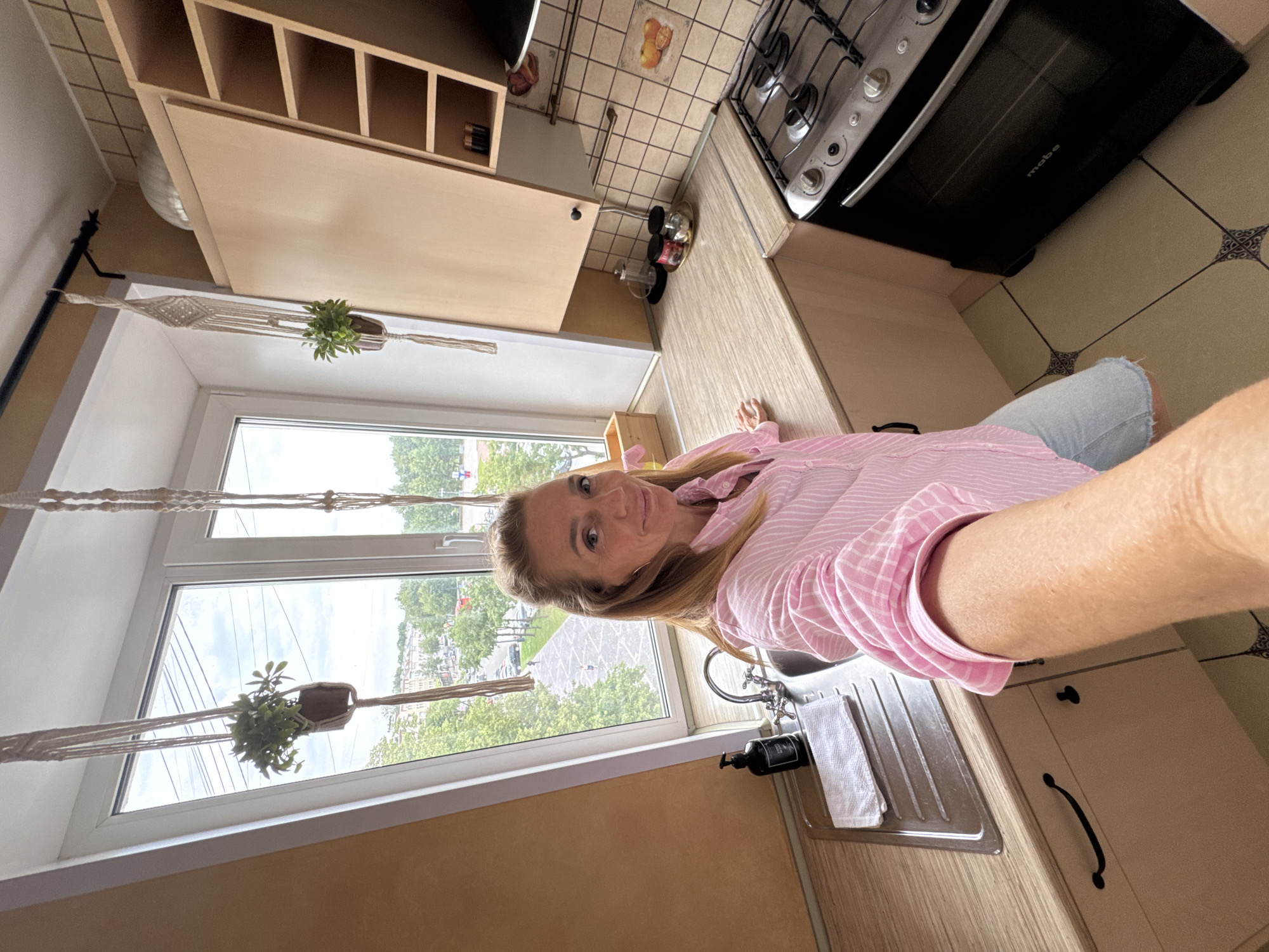 A woman is taking a selfie in a small kitchen. She is wearing a pink and white striped shirt and denim shorts. The kitchen has a sink, a stovetop, and cabinets. There are also two potted plants hanging in macrame hangers near the window. Outside the window, there is a view of a city street with trees and buildings. The scene appears to be in Veliky Novgorod, Russia.