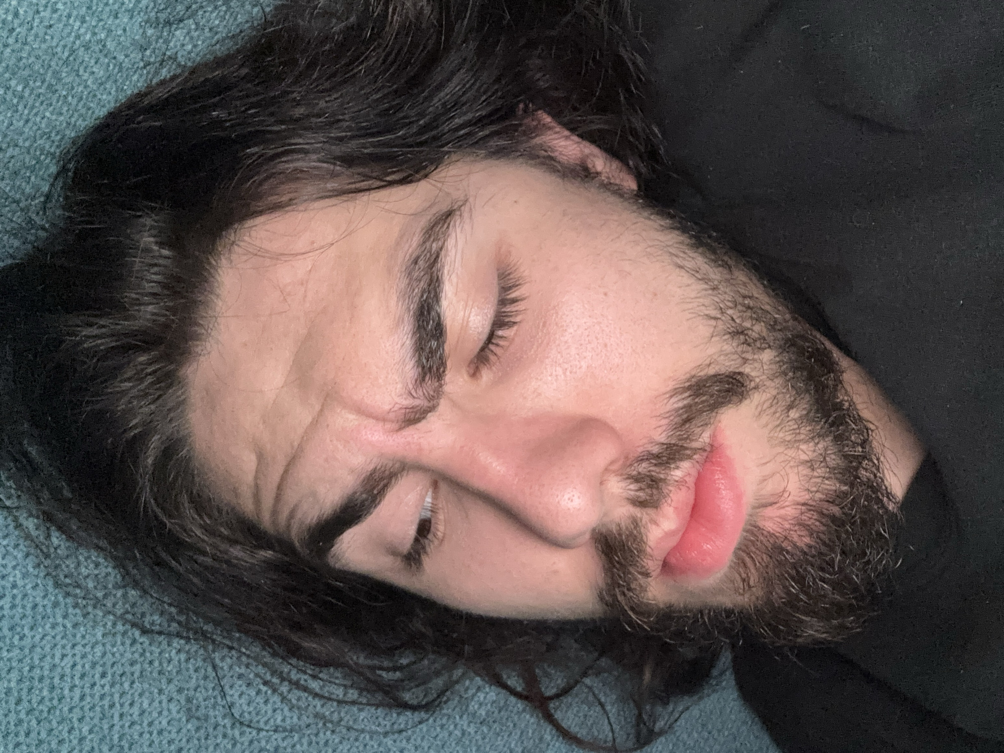 A close-up, overhead shot shows the face of a young man with dark, wavy hair and a beard. He is lying down, with his eyes closed and his mouth slightly parted. His expression is relaxed, almost sleepy. The background is a textured, teal-colored surface, possibly a blanket or sofa. He is wearing a black shirt.