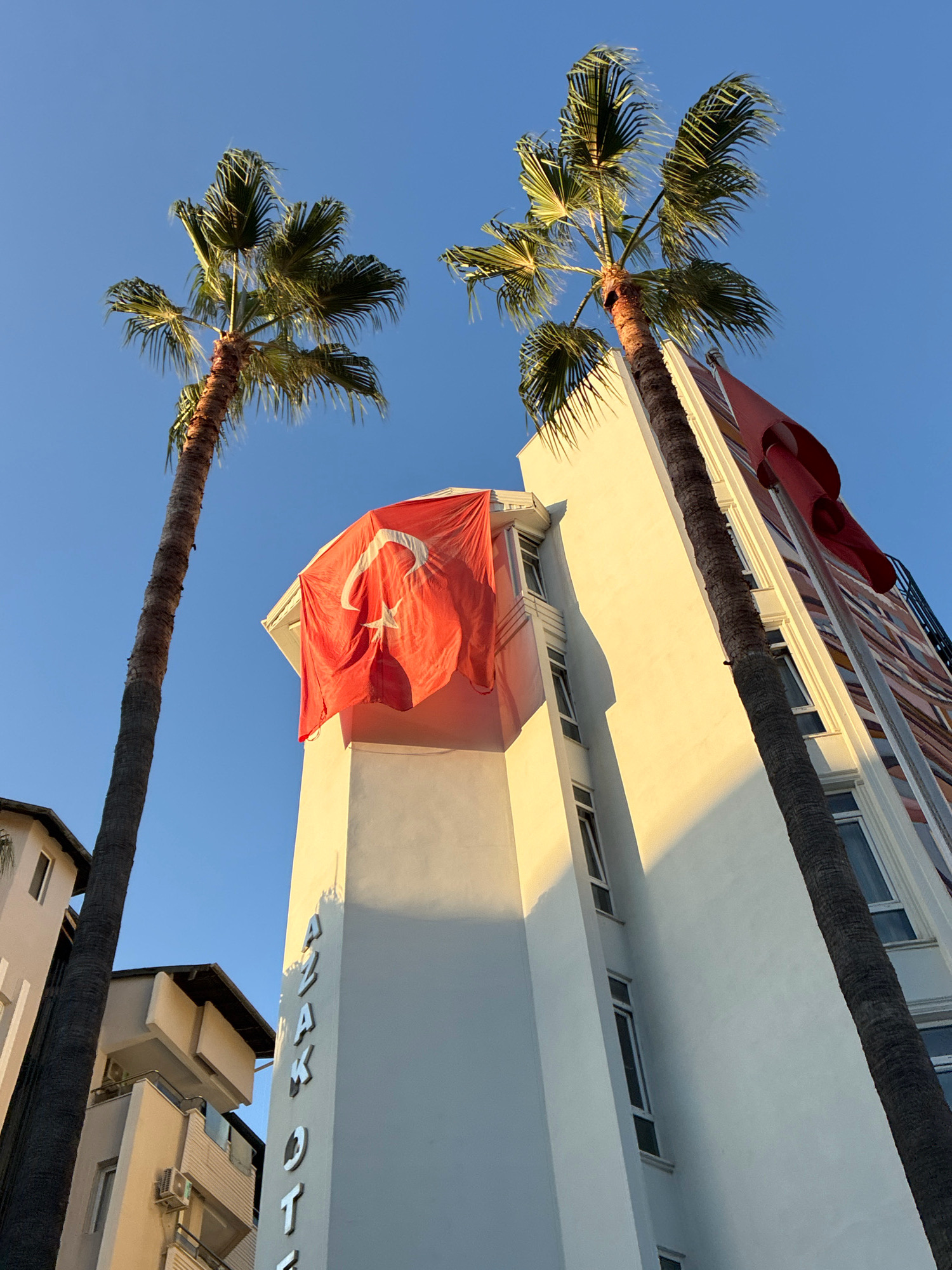 The image shows a low-angle view of a building in Alanya, Turkey, with a Turkish flag prominently displayed. Two palm trees frame the building, one on the left and one on the right. The building is white and has a modern design with many windows. The Turkish flag, a red flag with a white star and crescent, is draped over the side of the building, partially obscuring some of the windows. The sky is clear and blue, indicating it is daytime. The lighting suggests it is either morning or late afternoon, with the sun casting a warm glow on the building. The word "AZAK HOTEL" is visible in raised letters on the side of the building, identifying the location. Other buildings, including a darker, more colorful one in the background, are also visible, suggesting an urban setting. The image appears to be taken from street level, looking upwards.