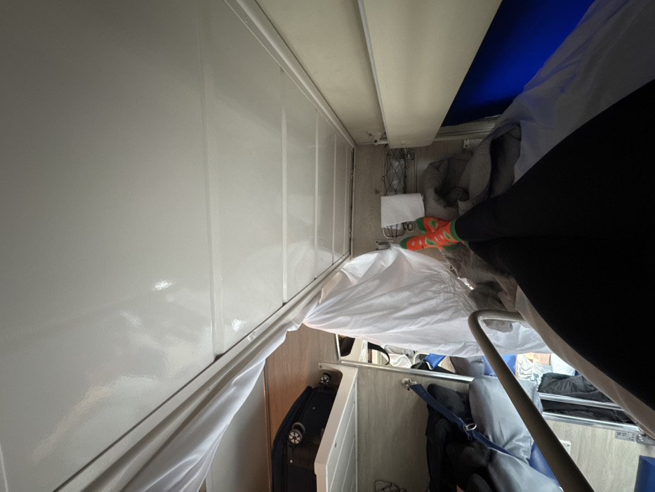 The scene is an interior view of a train sleeping carriage, consistent with *platskart* class coaches utilized in Russia, and is noted as potentially occurring in the vicinity of Gryazi, Russia. The perspective is from a lower bunk, looking towards the foot of the bed and the opposite side of the carriage.
In the immediate foreground, the legs and feet of an individual are visible, clad in black leggings and distinctive orange socks adorned with green and red patterns, with the feet crossed at the ankles. The individual rests on a bunk covered by white sheets and a grey blanket. To the immediate left, a white privacy curtain is partially drawn, suspended from a ceiling-mounted track. To the right, a white wall panel features a wire storage basket containing a folded white towel, positioned above a blue panel section.
The carriage ceiling is white and glossy. In the background on the left, a black wheeled suitcase with visible wheels is stowed on an upper bunk, and a lower bunk is partially visible with dark items. Metal safety rails are integrated into the bunk structures. The interior walls beyond the immediate bunk area are light brown.