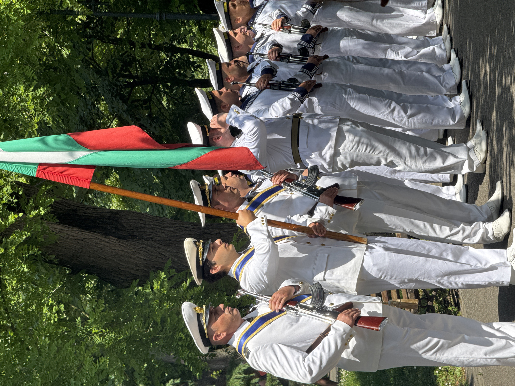 A group of male naval or military personnel, dressed in crisp white uniforms with distinctive white hats, are depicted in formation outdoors during what appears to be a parade or ceremonial event. There are at least seven men visible, standing in a line, many holding rifles that resemble AK-47 variants with wooden stocks. Some of the uniforms feature blue and gold trim or sashes.
At the front of the formation, one individual carries a flag on a wooden pole. The flag displays horizontal stripes of green, white, and red. Although the standard horizontal order for the Bulgarian flag is white, green, red, the colors present are consistent with the national flag of Bulgaria. This is further supported by visible text on some of the sailors' hats, which clearly reads "БЪЛГАРИЯ" (Bulgaria).
The setting is an outdoor area with abundant green foliage from trees and bushes in the background, suggesting a park or a tree-lined street. The ground beneath the men's white boots appears to be pavement or asphalt. The bright lighting and shadows indicate it is daytime, likely during clear, sunny weather. The men maintain a formal and disciplined posture, looking forward, typical of a ceremonial guard.