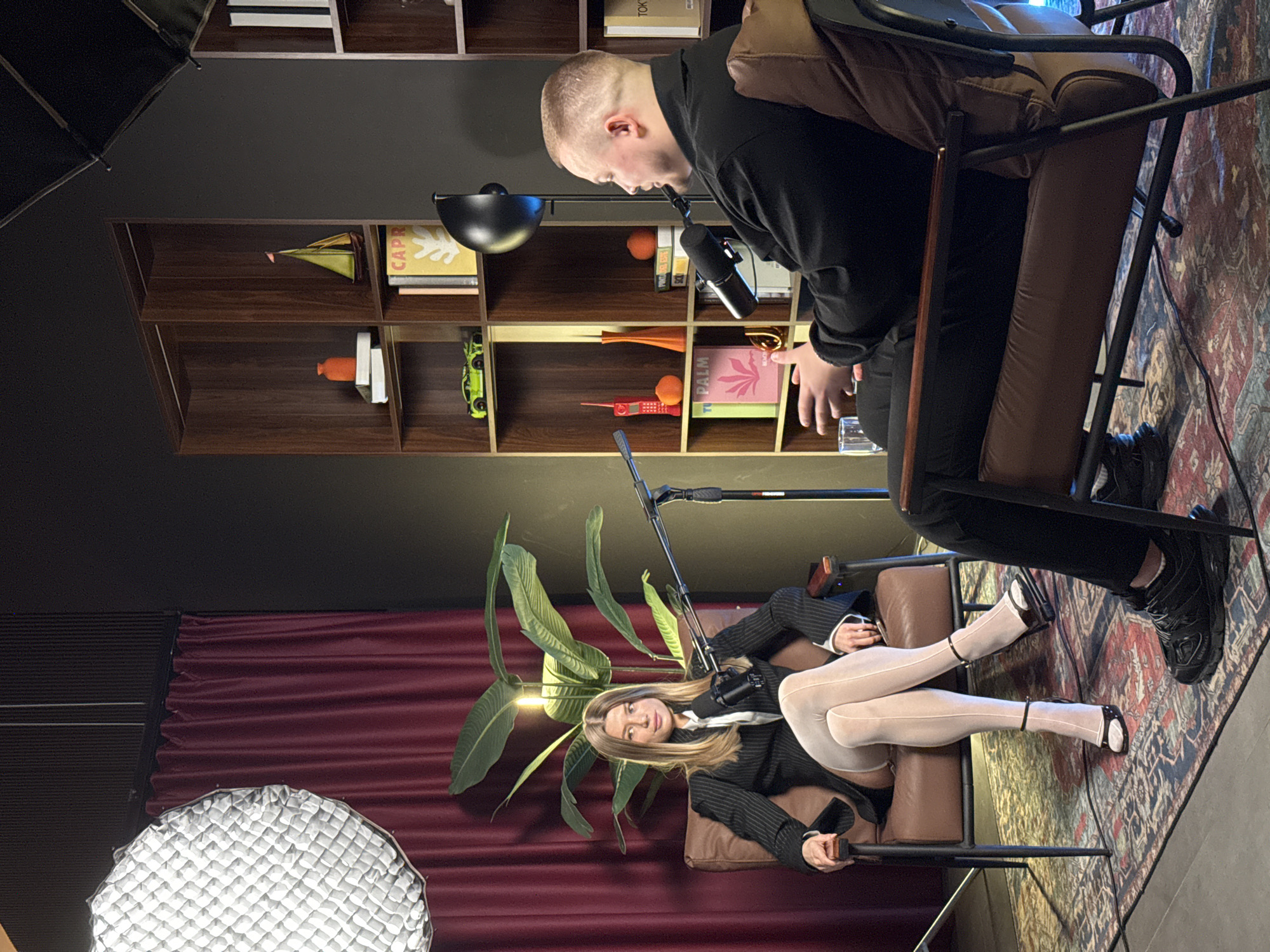 The image depicts a studio setting, likely in Warsaw, Poland, with two individuals engaged in what appears to be a podcast recording. A man with a shaved head is seated in a brown leather chair, leaning towards a microphone. He is wearing a black top and black pants, and black sneakers. Opposite him, a woman is also seated, dressed in a black and white striped suit, white stockings, and black and white sandals. She is seated in a similar leather chair. A microphone stands between them, indicating the recording setup.
Behind the man, there is a dark grey wall with a built-in bookshelf containing various objects, including books, decorative items, and toys. To the left of the woman, there's a burgundy curtain and a large softbox light. A tall green plant is positioned behind the woman, adding to the studio's ambiance. A patterned rug covers the floor, and additional lighting equipment is visible, contributing to the professional setting. The overall scene suggests a planned and staged recording environment.