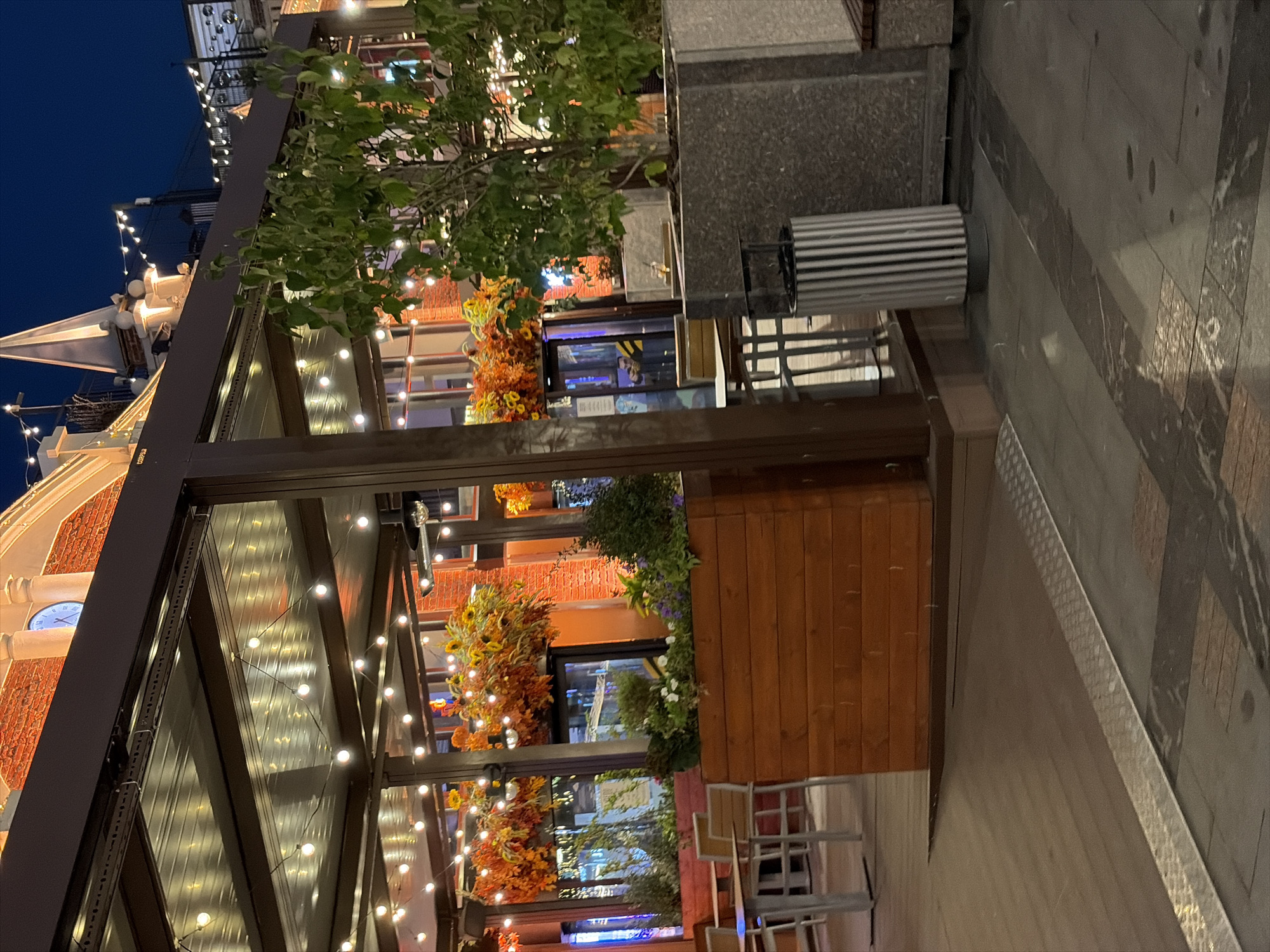 The image depicts an outdoor seating area of a restaurant or cafe at dusk, likely in a city environment. A covered patio with string lights illuminates the scene. In the background, a building with a brick facade and a clock tower is visible. The foreground shows a wooden planter box filled with flowers and greenery, and a trash can. The pavement is paved with large stone tiles and a metal strip. The overall impression is of a well-lit, inviting outdoor space. The specific location in Moscow, Russia, is not identifiable from the image alone.