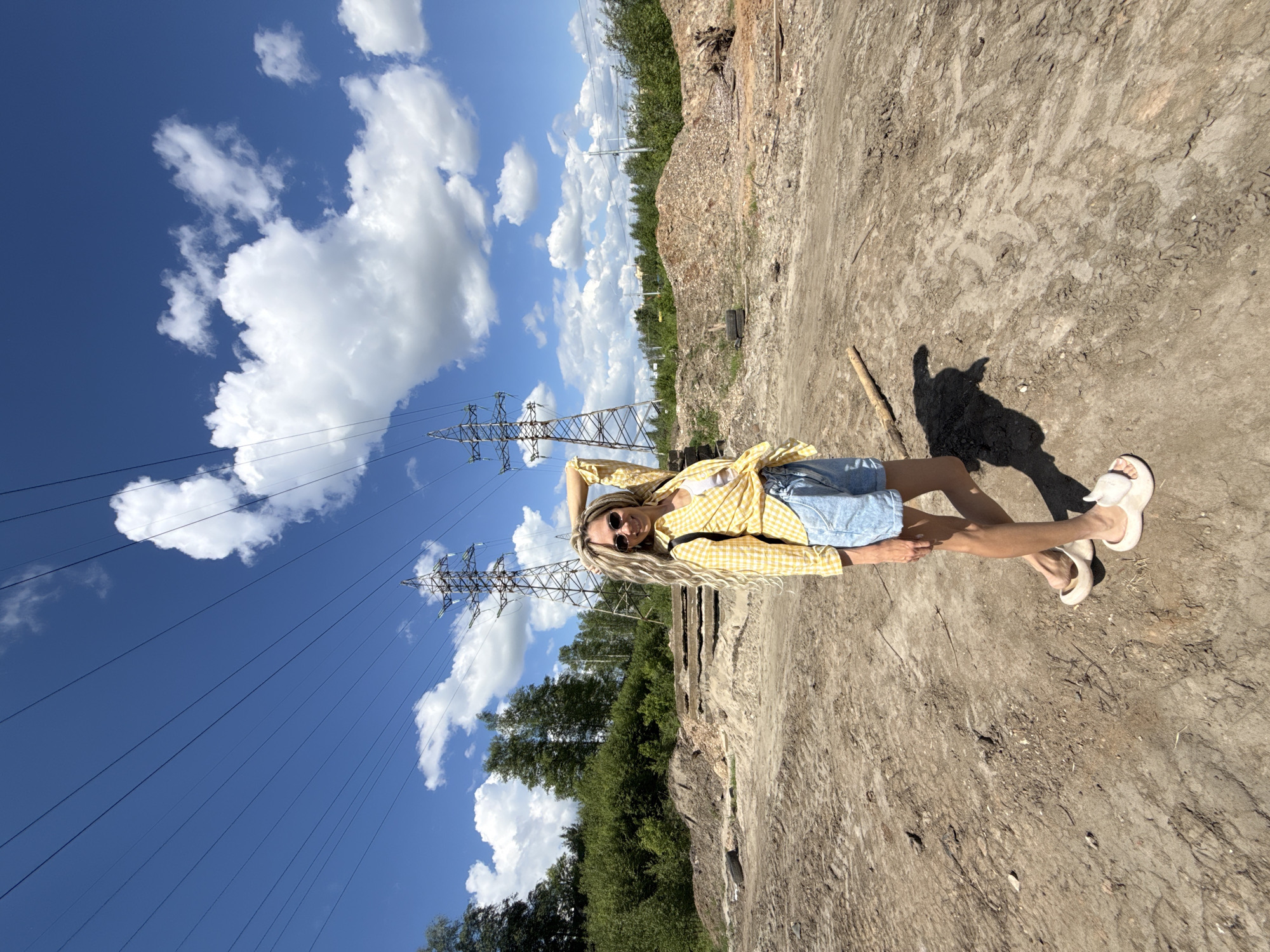 A young woman stands on dry, cracked soil in an outdoor setting in Заволжье, Russia. She wears a yellow plaid shirt, light blue denim shorts, sunglasses, and white sandals. She has long, wavy, light blonde hair. Her right hand is raised behind her head.
In the background, there are power lines and towers against a clear blue sky with white clouds. A tree line is visible behind the power lines and to the right of the woman. The ground appears to be an industrial or construction site. A shadow of the person and a stick are visible on the ground to the right.