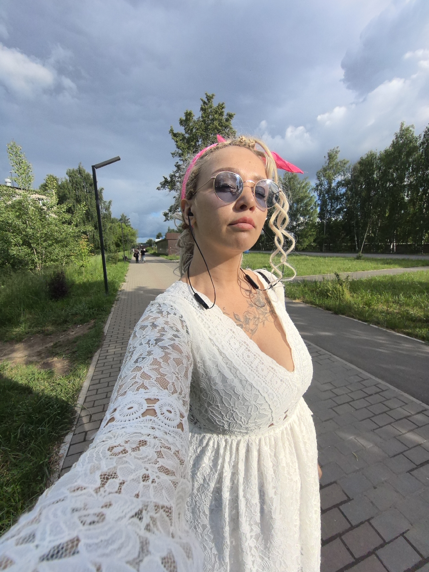 A woman with blonde hair styled in braids and a pink bandana takes a selfie while walking on a paved path in Chaikovsky, Russia. She is wearing a white lace dress and sunglasses, with black earbuds in. The path is lined with green grass and trees, and a lamppost is visible in the background. The sky is cloudy.