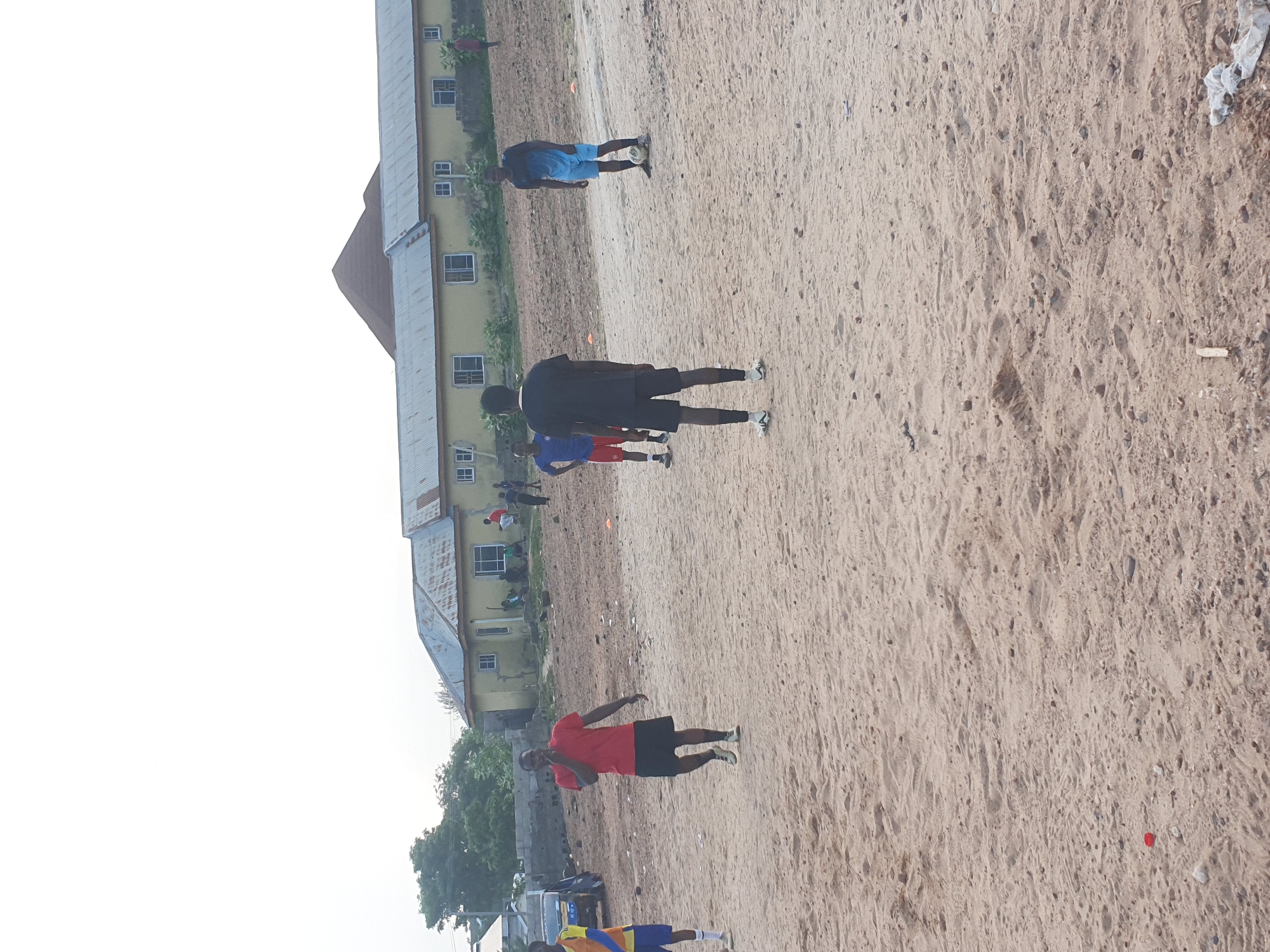 The image depicts a sandy open area in Lokoja, Nigeria, where several individuals are engaged in physical activity. A long, two-story building with a light-colored facade and a gray roof runs along one edge of the open space. People are seen wearing various colored shirts and shorts, some appear to be jogging or walking. Small orange cones are sporadically placed on the sand. The sky appears overcast. Some vegetation is visible near the building.