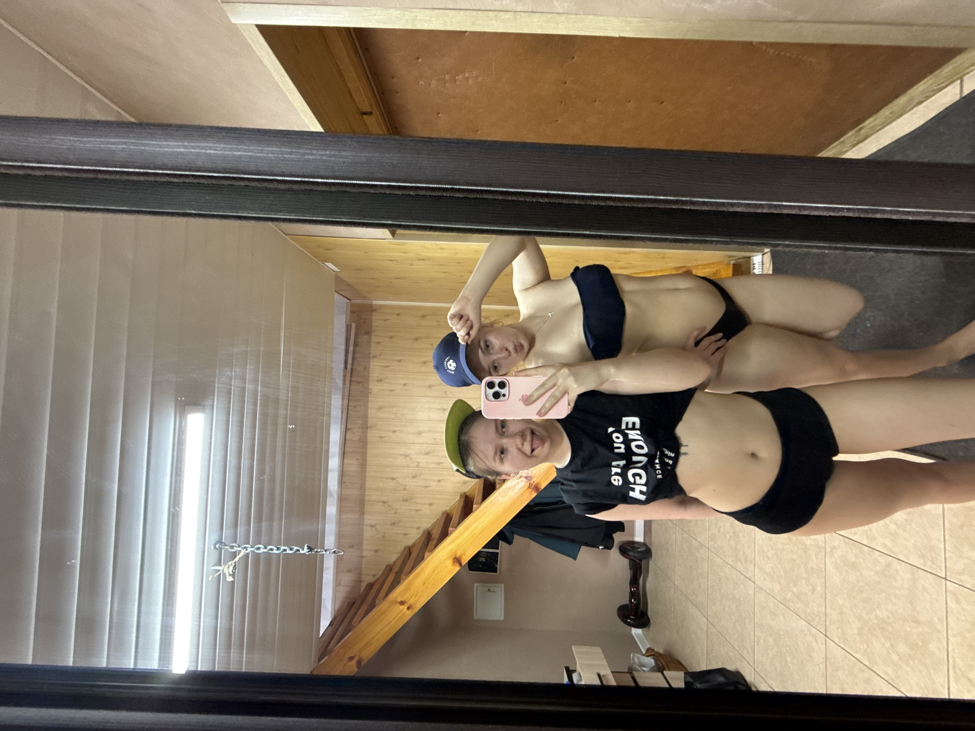 Two women are reflected in a mirror. One woman wears a blue baseball cap and a dark two-piece swimsuit. The other woman wears a green baseball cap, a black t-shirt with white lettering, and black underwear. The reflection shows a room with wooden walls, a wooden staircase, a hoverboard, and tiled flooring. The location is likely Zhigalki, Russia, based on the image request.