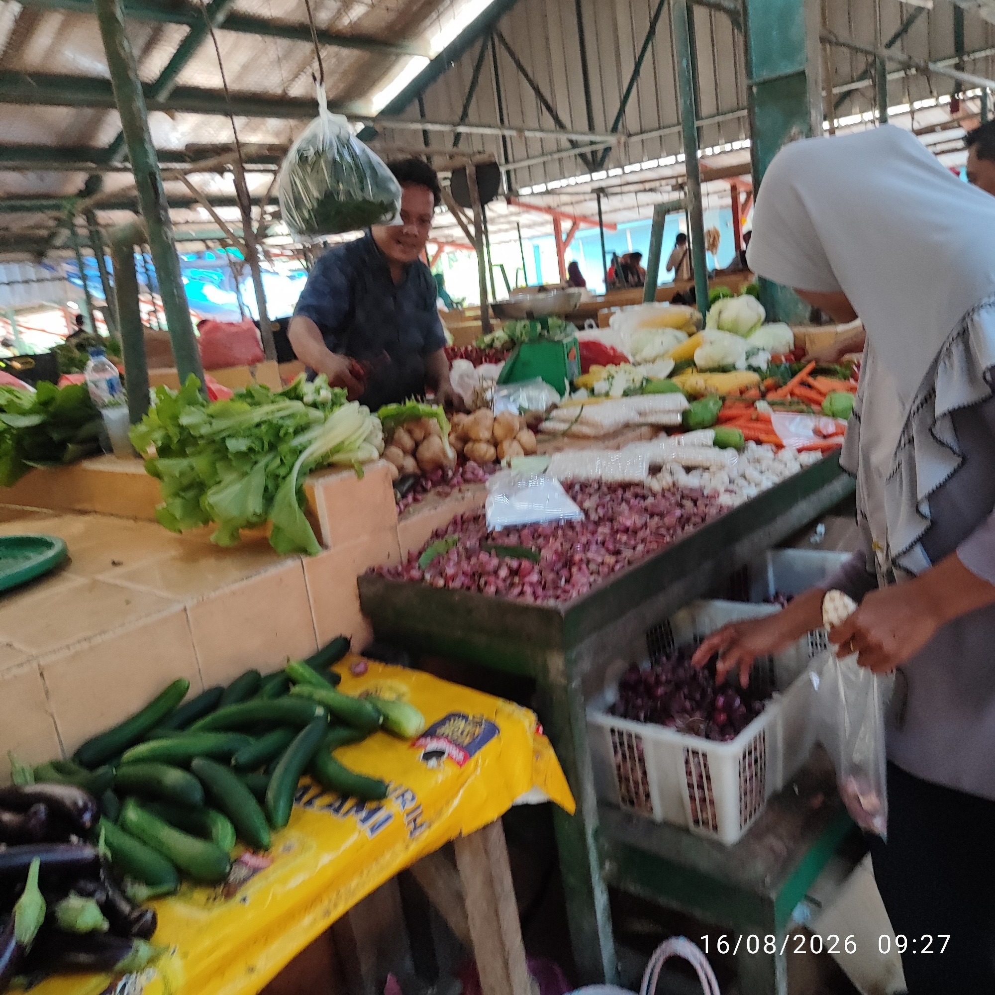 The image depicts a bustling outdoor market scene in Pembaharuan, Indonesia, likely in the morning given the timestamp "16/08/2026 09:27". A man in a dark blue shirt is actively engaged in selling produce. He stands behind a stall laden with various vegetables, including leafy greens, onions, potatoes, and what appear to be cucumbers and eggplants. A large plastic bag filled with leafy vegetables hangs overhead. To the right, a woman wearing a grey hijab and a grey top is interacting with the stall, possibly purchasing items. She holds a plastic bag and appears to be selecting produce from a bin of red onions. The market environment is characterized by open-air stalls with overhead roofing supported by metal and bamboo structures. The ground appears to be tiled, and the overall impression is one of a vibrant, local marketplace.