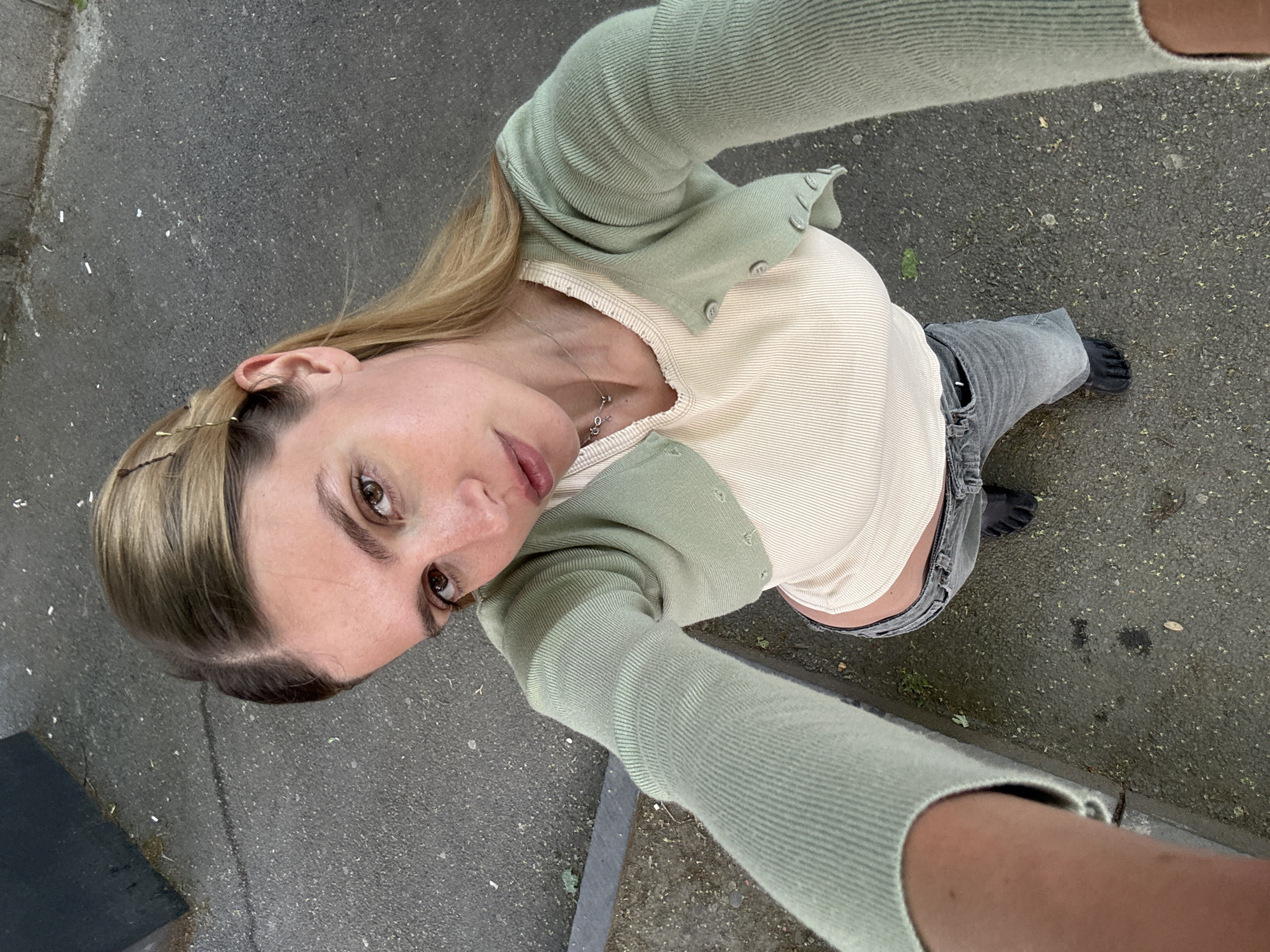 A woman stands on gray asphalt, angled to the left and looking up at the camera. She has blonde hair with dark roots and is wearing a light green cardigan over a beige top, with gray jeans and black footwear. A silver necklace is visible around her neck. The background shows a textured, gray asphalt surface, possibly a sidewalk or street in Tbilisi, Georgia, as indicated by the prompt. The angle suggests the photo was taken in a selfie-style, with the woman holding the camera above her. A gray curb is visible along the bottom edge.