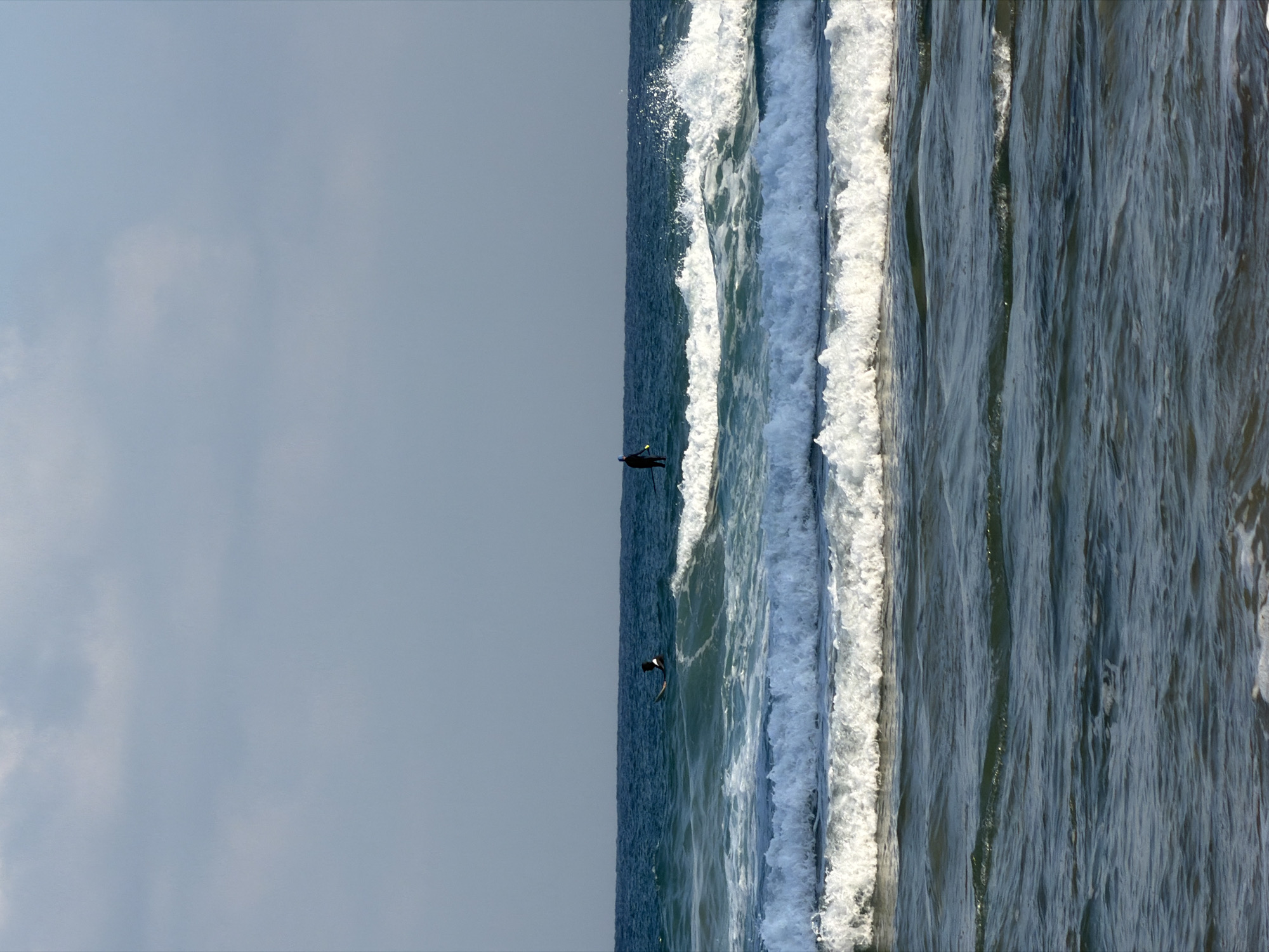 This is a photograph of the Mediterranean Sea in Bat Yam, Israel. In the distance, two individuals on surfboards are visible. One person is sitting on their board, and the other is standing. Both are facing away from the camera. The water is choppy with several waves breaking, creating white foam. The sky is a pale blue with some scattered clouds. The photograph is taken from a low angle, with the water filling the majority of the frame. The sun appears to be behind the camera, casting a bright light on the scene.