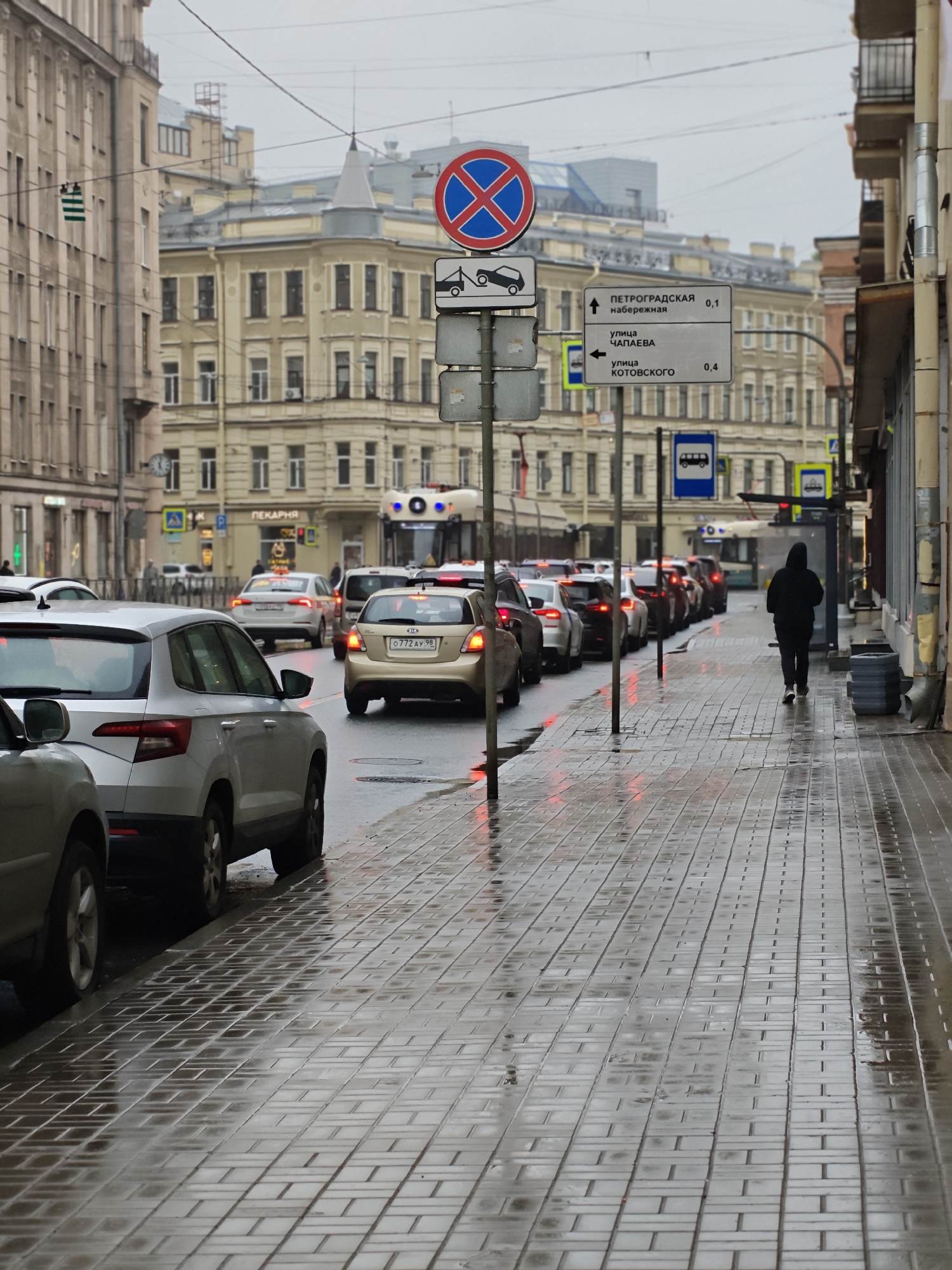 This is a street-level, medium-shot photograph taken on a cloudy, overcast day, likely in the morning or late afternoon. The wet, reflective cobblestone sidewalk and the gleaming wet street suggest recent or ongoing rain. A line of cars is stuck in traffic on the right side of the street, with their brake lights illuminated. On the left, the sidewalk is lined with parked cars.
The scene is an urban setting with multi-story buildings lining the street. The architecture of the buildings suggests an older European city, possibly Eastern Europe, with ornate facades. A prominent sign in the upper left quadrant reads "ПЕКАРНЯ," which translates to "Bakery" in Russian.
Several traffic signs are visible, including a "No Parking" sign and directional signs with place names and distances in Russian. One sign indicates "ПЕТРОГРАДСКАЯ набережная" (Petrogradskaya Embankment) with a distance of 0.1, and another points to "улица ЧАПАЕВА" (Chapaev Street) and "улица КОТОВСКОГО" (Kotovsky Street) with a distance of 0.4. A bus is also visible further down the street.
A lone figure, dressed in dark clothing and a hooded jacket, walks away from the camera down the sidewalk on the right. Their posture suggests a solitary, perhaps hurried, movement. The overall mood of the image is somber and mundane, conveying the routine of urban life amidst inclement weather. The reflections on the wet surfaces add a touch of visual interest to the otherwise gray scene.