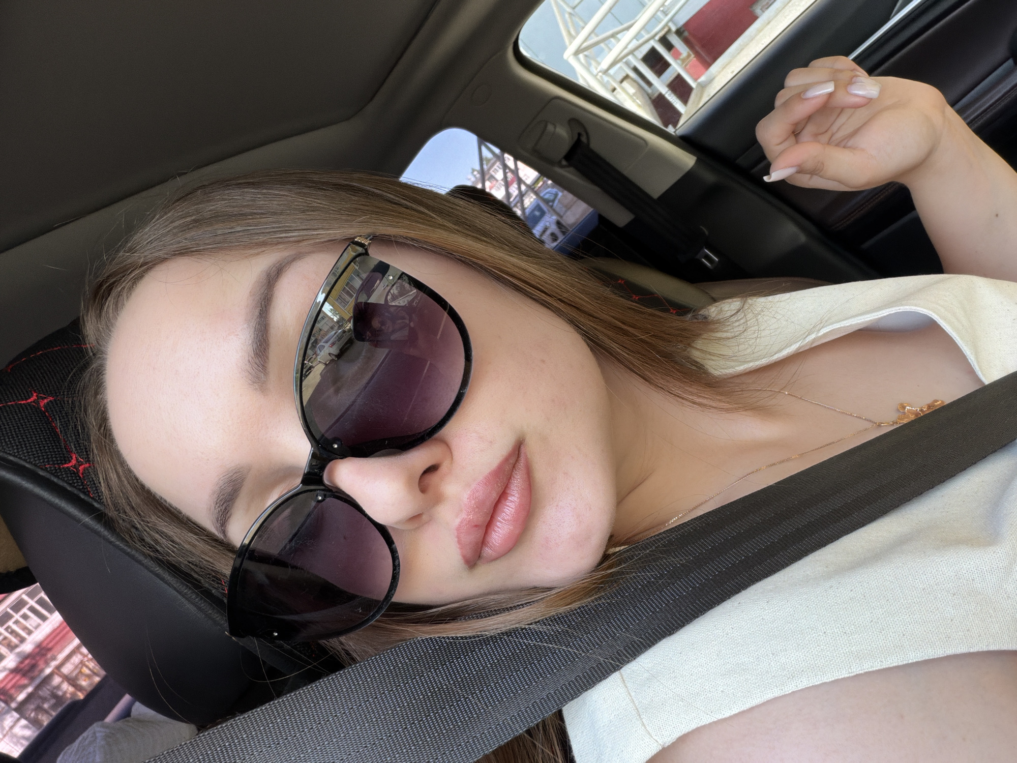 A young woman is photographed from a slightly upward angle, positioned inside a car, likely in the passenger seat. She is wearing dark sunglasses and a white top, with a black seatbelt crossing her torso. She is wearing a gold necklace. Her hand is resting on her upper arm. The car's interior is visible in the background, along with glimpses of the outside environment, including buildings and what appears to be metal scaffolding. The reflection in her sunglasses suggests the location is the city of Chaikovsky, Russia.