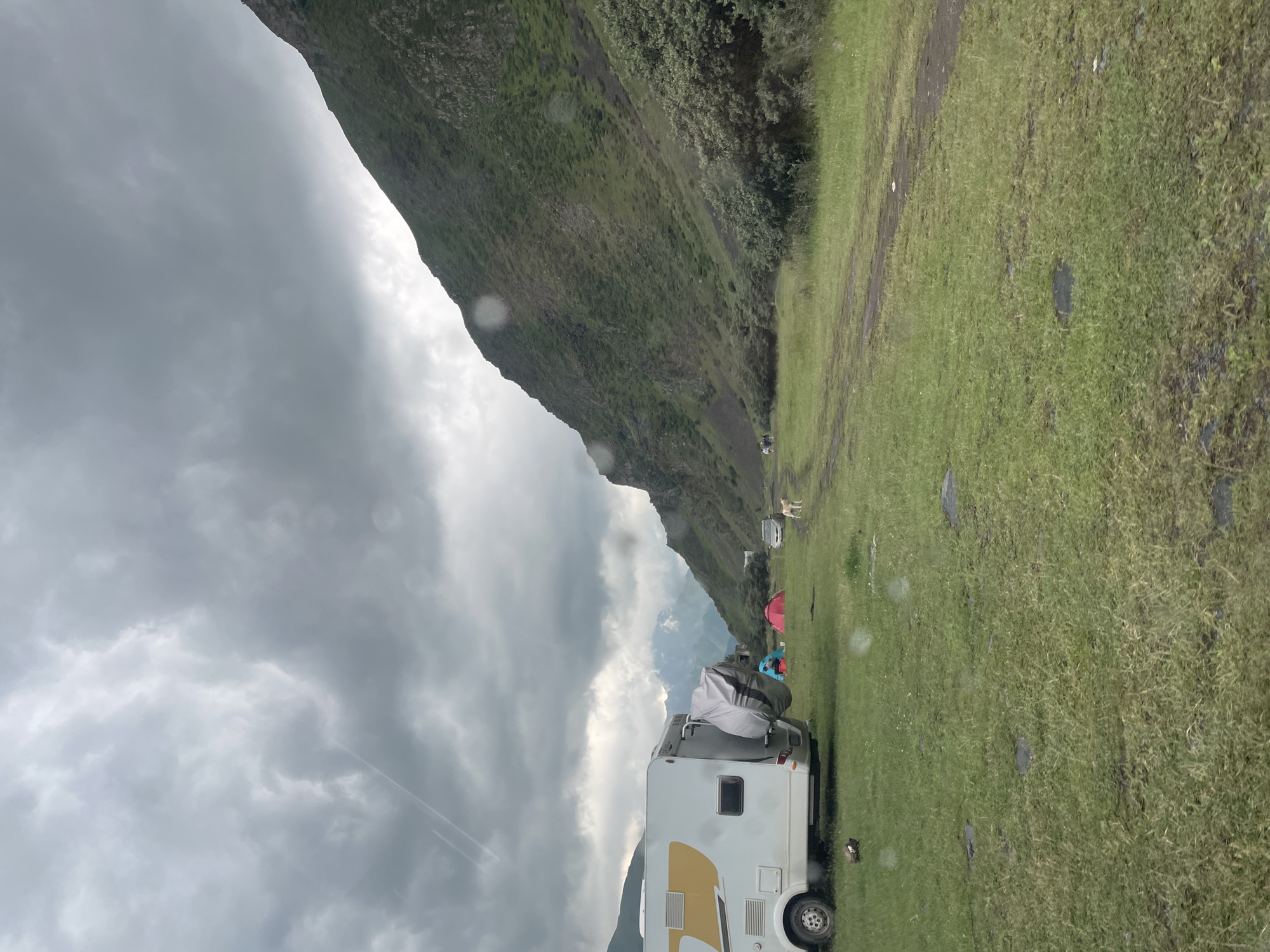 A cloudy day scene depicts a campsite in a valley near Stepantsminda, Georgia. In the foreground, a white and yellow camper van is parked on a grassy area. To the left of the camper van, a spare tire cover is visible. Behind the camper van, there are two tents, one red and one blue. A white car is parked further back in the scene, and a light-colored dog stands near it. The terrain consists of a dirt path leading into the distance, with grassy fields on either side. A steep, green, and rocky mountain slopes upwards on the right side of the image. In the background, distant mountains are visible, partially obscured by clouds. There are a few people visible near the distant mountains on the left side of the image. The sky is overcast with dramatic cloud formations.