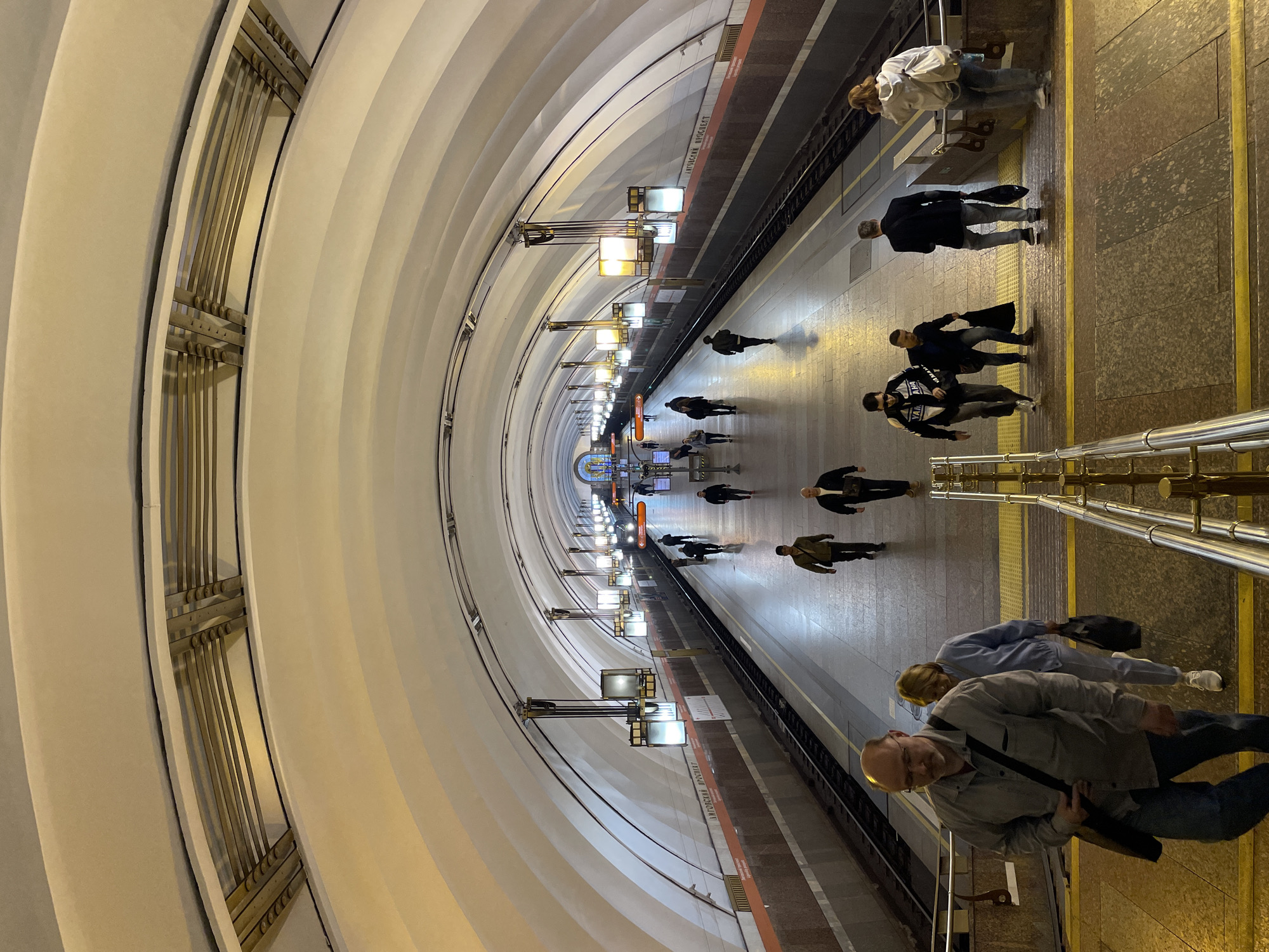 A nearly empty underground metro station platform is captured in a perspective that accentuates the arched architecture of the roof and ceiling. The walls curve inward and are adorned with vertical metal bars. Below, a polished stone floor forms the platform, where a few people are scattered, some walking in pairs or small groups. Overhead lamps hang at regular intervals, casting a soft, ambient light. The scene is devoid of trains, adding to the sense of emptiness. In the foreground is a man, with his back facing the viewer, positioned near a set of stairs, adding a human element to the scene. The picture is taken in Moscow, Russia at the station Arbatskaya.