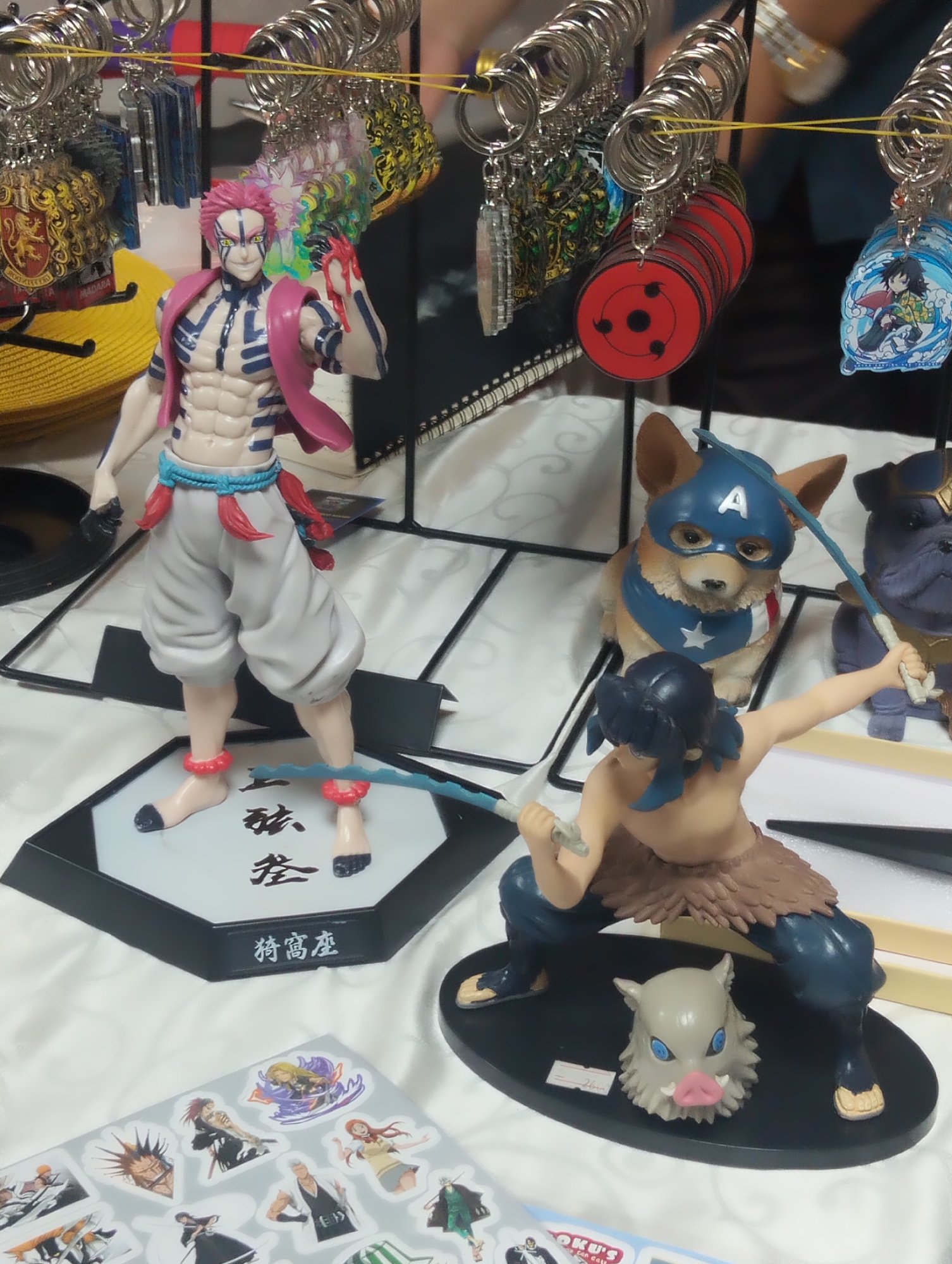 This image displays a collection of anime-themed merchandise, likely at a convention or market stall. In the foreground, several figures are arranged on a white cloth surface. The most prominent is a male anime figure with pink hair, a muscular physique, and light gray pants, holding a blue sword. To the right of this figure is another smaller figure of a boar's head. Further back, a corgi dog wearing a Captain America mask and cape sits next to a dark-colored dog figure that appears to be Thanos. Behind these figures, a display rack holds various keychains featuring anime characters and symbols. The background is blurred but suggests a crowded indoor setting with other displays and possibly people. There is no indication of the specific city of Dhaka, Bangladesh, in the image itself.