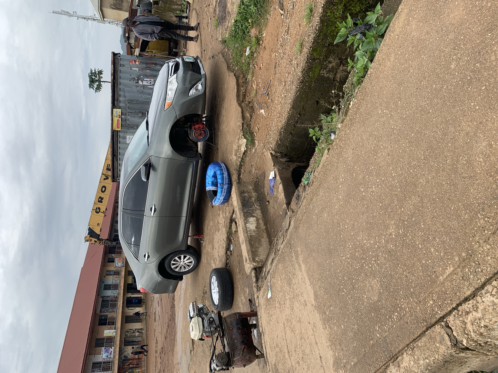 The image depicts an outdoor scene in Rafin Par, Nigeria, likely a makeshift tire repair shop. A grey sedan is positioned with its front passenger-side wheel removed and supported by a jack. A spare tire with a blue cover is placed on the ground nearby. To the left of the car, a red air compressor with a white engine sits on a stand, alongside another tire. A man in a grey jacket and dark pants stands to the right of the car, observing the scene. In the background, a multi-story building with a red roof is visible, along with shipping containers and a yellow crane. The ground is uneven and appears to be dirt and concrete, with a drainage ditch running along the bottom edge of the image. Overcast skies suggest it may be a cloudy day.