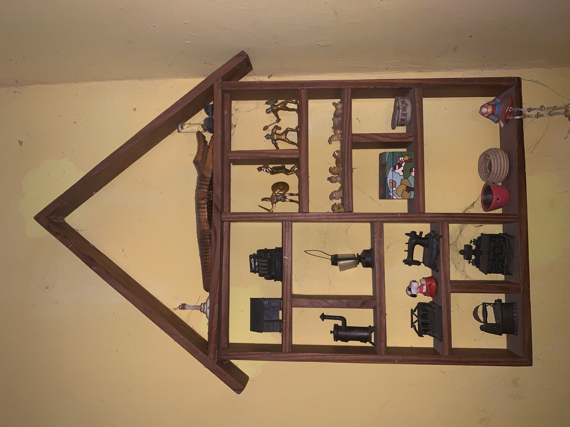 The image shows a wooden house-shaped shelf mounted on a yellow wall. The shelf contains various small objects, including figurines of soldiers, teddy bears, a painting, decorative rings, miniature cast-iron stoves, and other metal objects. Some objects are also hanging off the shelf. The image was taken at a tilted angle. There is no definitive information about its location in Santa Ana, Venezuela.