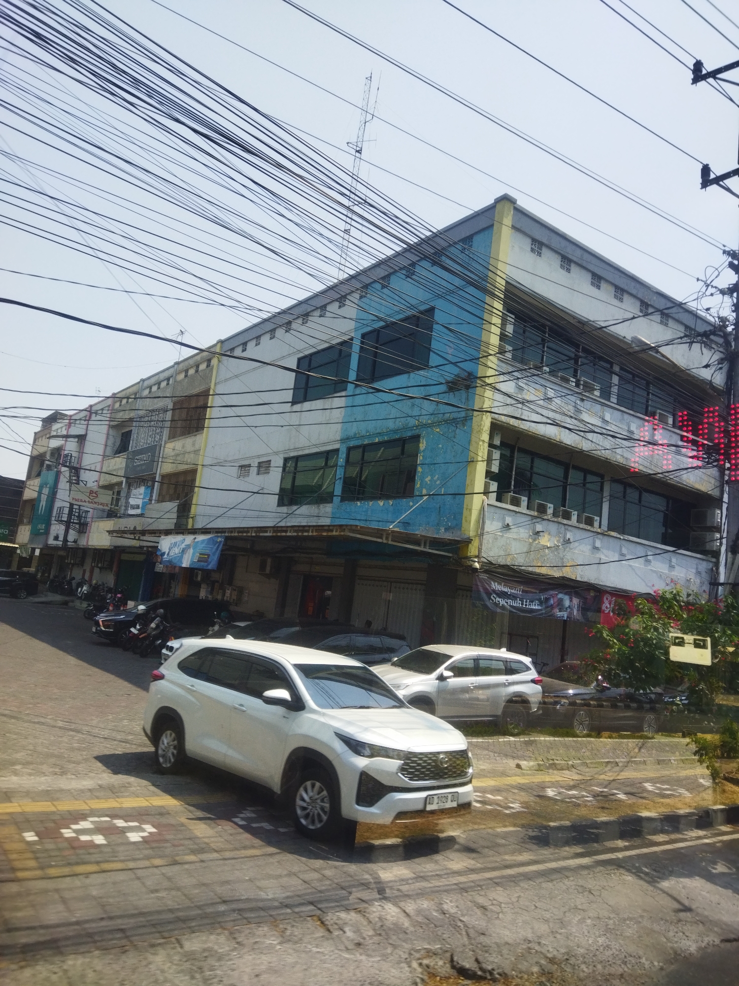 The image depicts a street scene in Grogol, Indonesia, dominated by a multi-story building with a blue and yellow facade. The building features numerous windows, many equipped with air conditioning units. A white SUV is parked in the foreground on the street. Several motorcycles are parked along the side of the street in front of the building. Overhead, a dense network of power lines crisscrosses the sky. A banner with Indonesian text is visible on the building. The street is paved and shows signs of wear. The sky is bright and clear, suggesting a daytime setting.