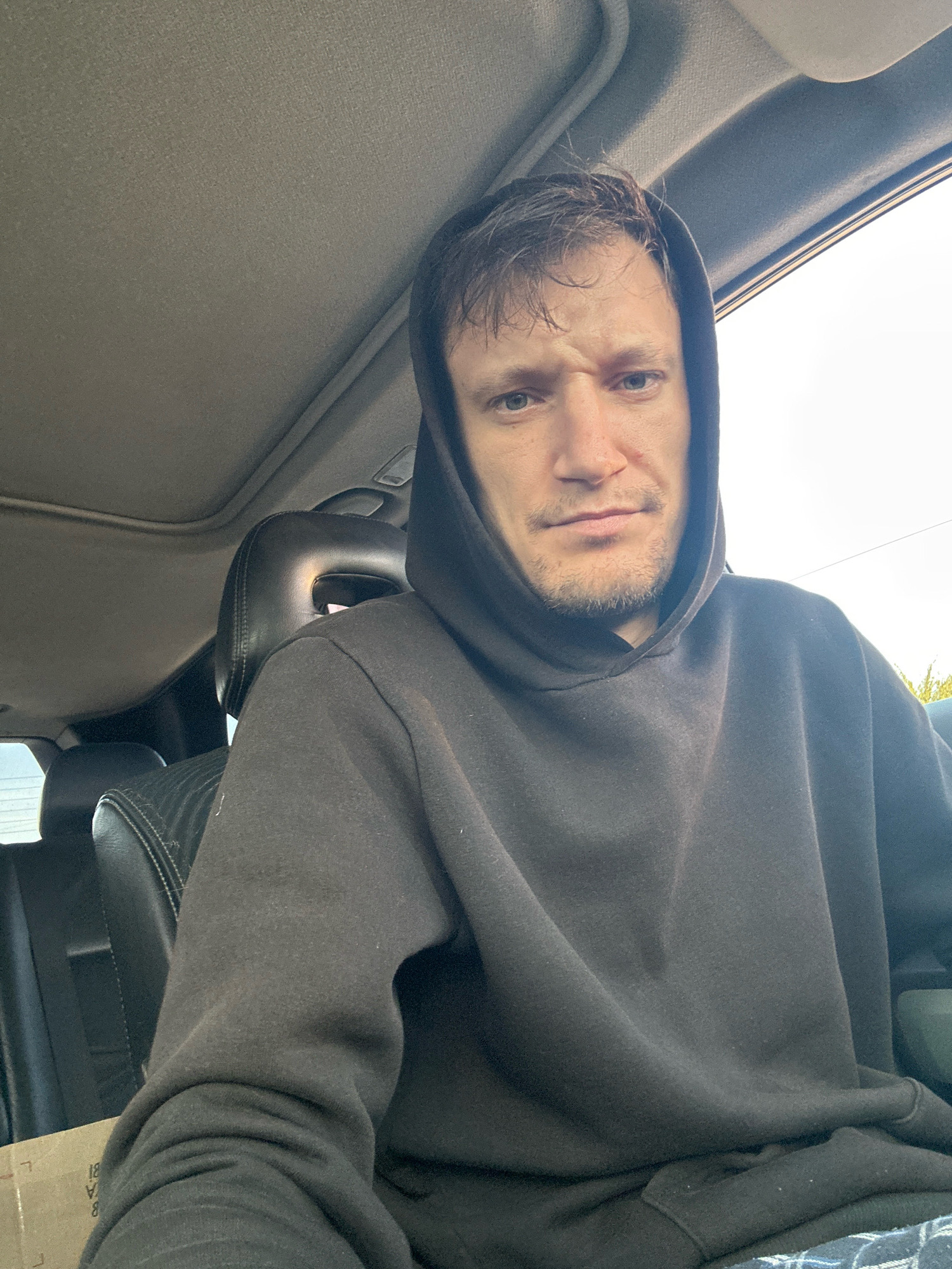A man in his early 30s wearing a dark grey hooded sweatshirt is sitting in the driver's seat of a car, looking directly at the camera. His hood is pulled up. The interior of the car is visible, including the headrest of the passenger seat and a portion of the sunroof. The man has short brown hair and stubble. The lighting suggests it is daytime. The location is Makiv, Ukraine.