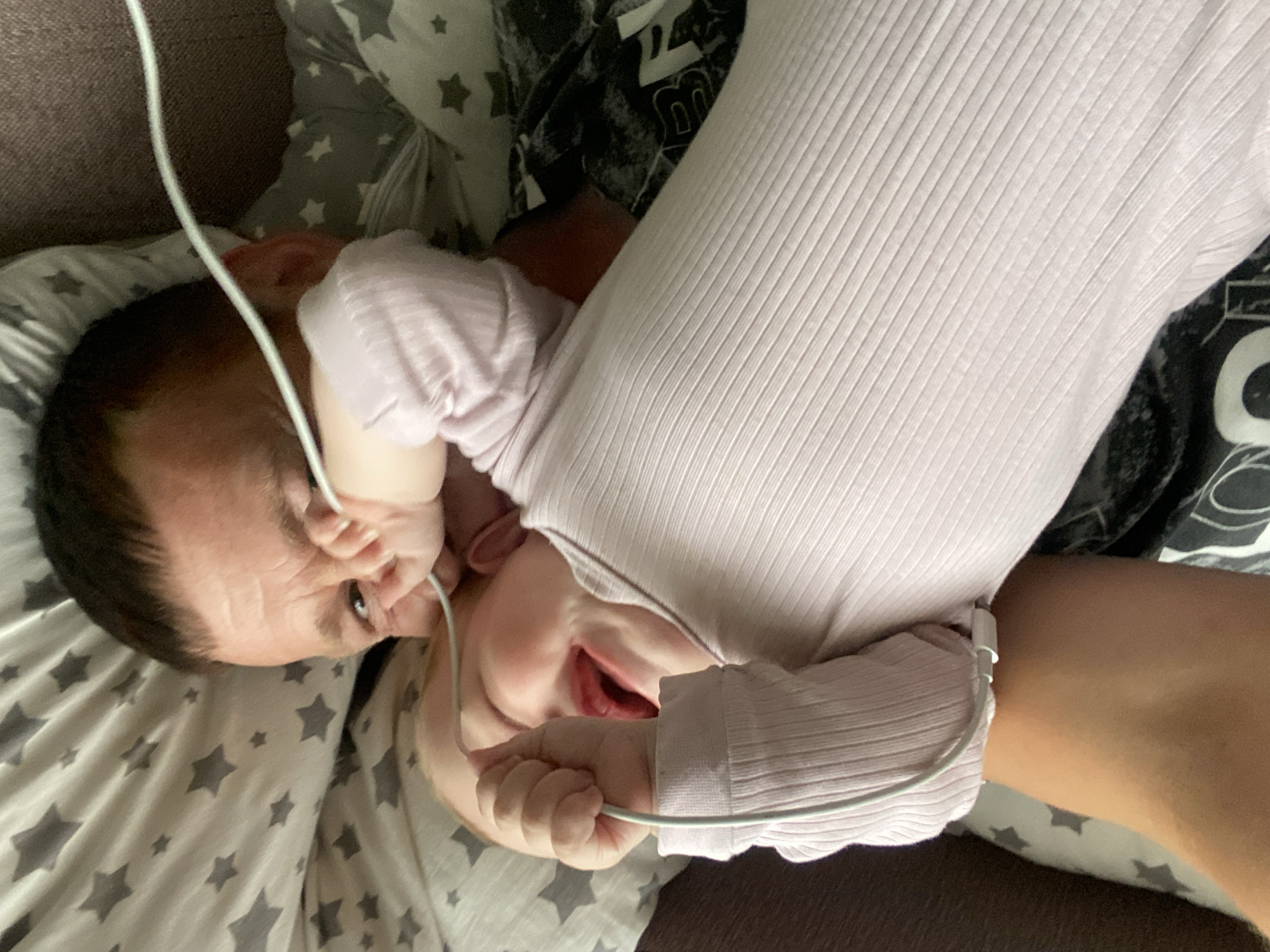The image depicts an infant and an adult lying together, likely on a sofa or bed, captured from an overhead perspective with the subjects oriented on their sides. The infant, with dark hair and wearing a light pink ribbed long-sleeved top, is the central focus. It is actively holding a white electronic cable in both hands, bringing it close to its face, with its mouth slightly open, suggesting curiosity or an attempt to interact with the object.
A portion of an adult's arm and torso, dressed in a light-colored ribbed garment similar to the baby's, is visible beside the infant. The subjects are surrounded by bedding, including a grey and white star-patterned pillow. Behind the adult, a dark patterned fabric with some indistinct white lettering is visible, but the text is illegible. The background consists of a dark, textured fabric, possibly upholstery.
The scene portrays a candid, intimate moment in an indoor, domestic setting. The lighting suggests daytime, though no specific time or outdoor conditions are discernible. The baby appears focused on the cable, but no strong emotions like joy or distress are clearly depicted.