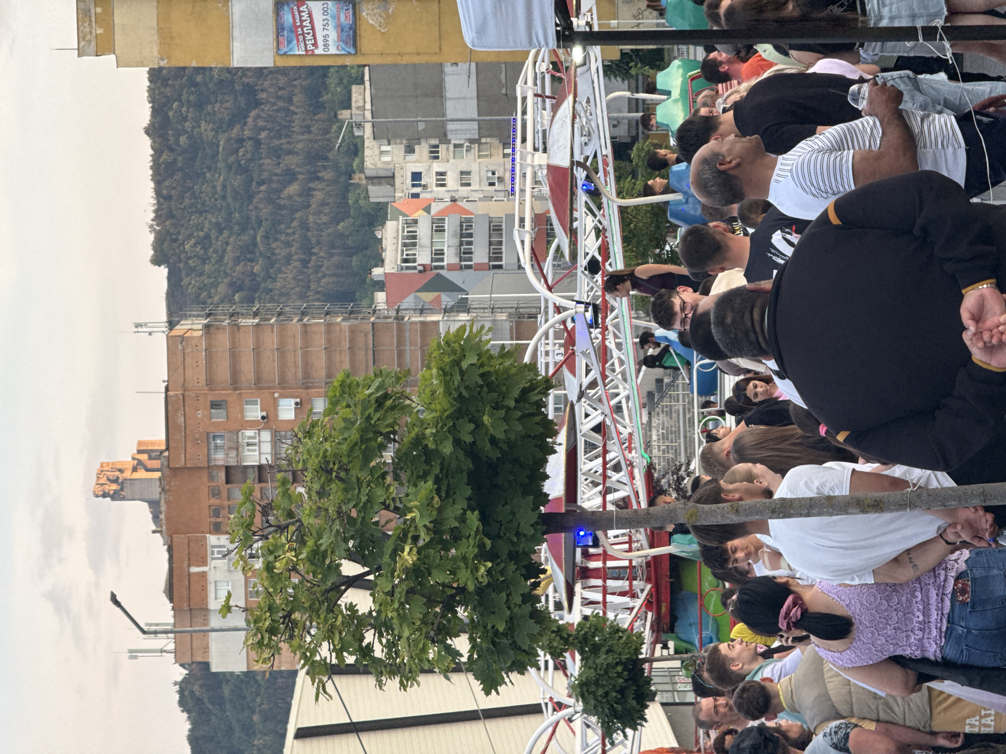 This image depicts a lively outdoor scene in Shumen, Bulgaria, featuring a large crowd gathered around an amusement park ride. The perspective places the viewer amidst the people, looking towards the ride and beyond to urban structures and a natural landscape.
In the foreground, a diverse group of people, mostly seen from behind or in profile, are casually dressed and appear to be observing or waiting. Dominating the right side of the frame is a large, white and red metal amusement park ride structure, resembling a compact Ferris wheel or a similar rotating attraction, with some people visible near or on its components. A large tree with vibrant green foliage partially obscures the ride in the lower foreground.
The midground features a collection of multi-story buildings with varied architectural styles. One building on the left has a light yellow facade and an advertising billboard. Another building in the center displays a distinct, colorful roof pattern with orange, blue, and green sections. Further back, buildings appear to be under construction or renovation.
The background is characterized by a densely forested hillside, which rises behind the urban area. The sky above is overcast and bright, suggesting daytime, possibly late afternoon or early evening.
The billboard on the yellow building clearly displays text in Bulgarian: "МЯСТО ЗА РЕКЛАМА" (Place for advertisement), "РЕКЛАМА" (Advertisement), and a phone number "0895 753 003". The overall atmosphere suggests a public gathering or fair.