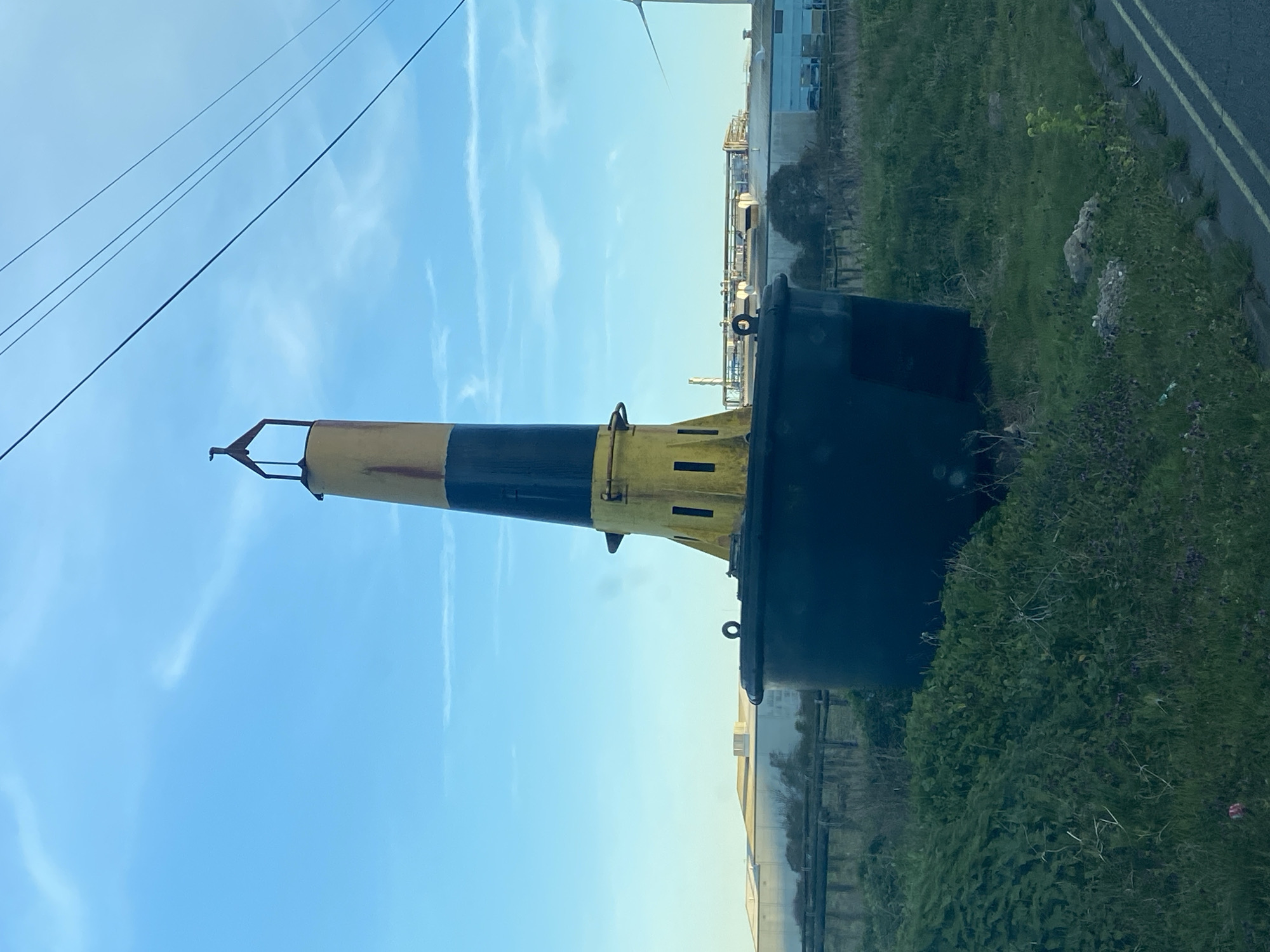 An inert, black and yellow navigational buoy lies on its side amidst roadside vegetation. The buoy, thick and tapered, is painted with horizontal bands of yellow and black. It is situated on a grassy verge beside a road, discernible by double yellow lines. In the background, there's an industrial complex and a clear blue sky with some light cloud cover. The location is likely in East Suffolk, United Kingdom, based on the landscape and infrastructure. Overhead electrical wires are also visible in the sky.