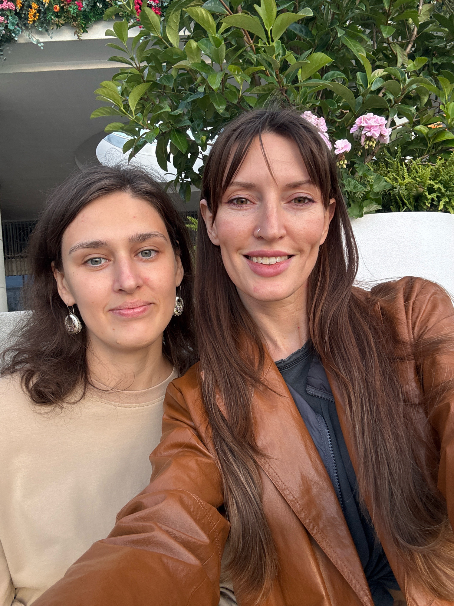 This is a close-up, waist-up, selfie-style photograph of two women, likely friends or family members, taken outdoors. The woman on the left has light skin, dark brown wavy hair, and is wearing a tan sweatshirt and large silver earrings. The woman on the right has light skin, long dark brown hair, and is wearing a brown leather jacket over a dark grey zippered top. She is also wearing a small nose ring. They are positioned side-by-side, with the woman on the right leaning slightly towards the woman on the left, and both are smiling at the camera.
Behind them, a lush green leafy plant with some small pink flowers is visible, suggesting an outdoor setting with vegetation. In the background, a portion of a light-colored building with architectural details like curves and a circular opening can be seen, along with some additional greenery. The lighting suggests it is daytime. The scene is clean and well-lit.
This image does not contain any elements that would suggest a specific location in Moscow, Russia.