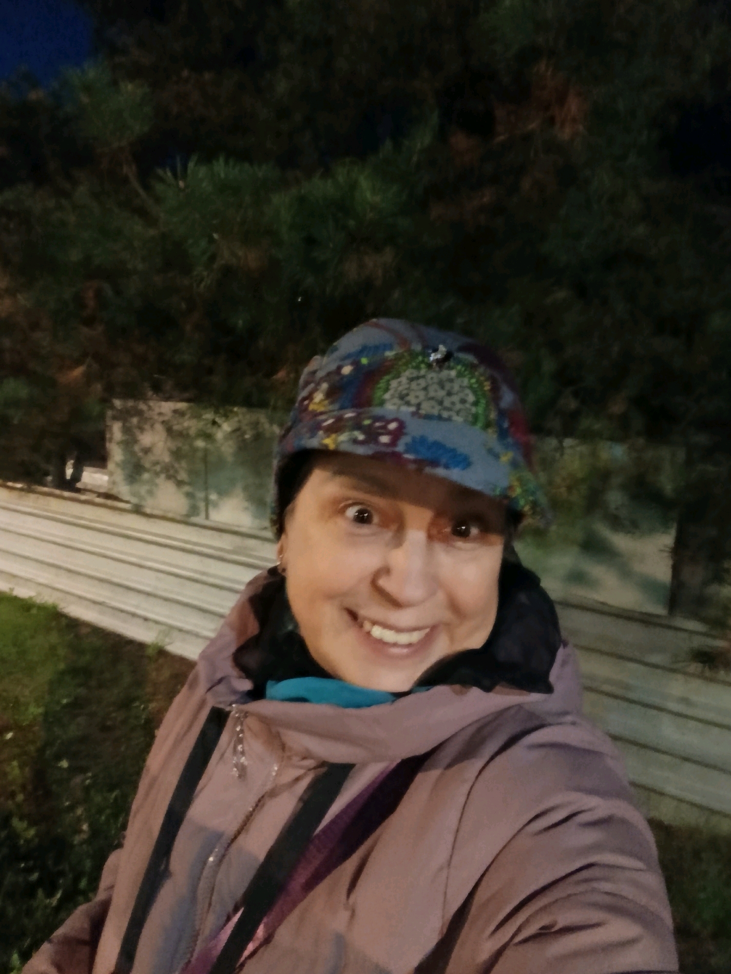 A woman wearing a patterned hat and a light brown jacket stands outdoors at night. The background shows dark foliage and what appears to be a concrete wall. The woman is smiling directly at the camera. The overall scene is dimly lit, suggesting it is evening or night. The location is specified as Verea, Russia.