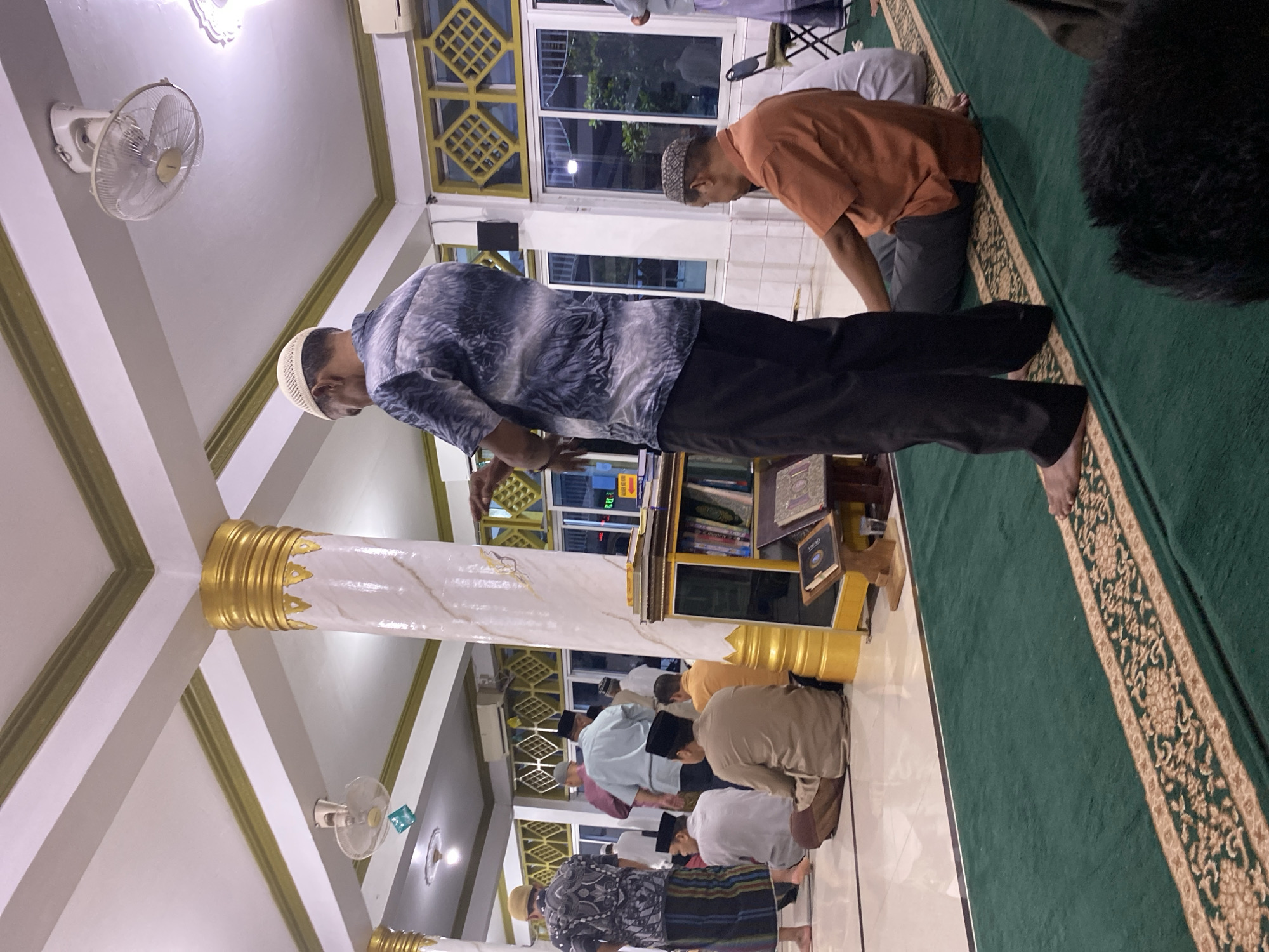 The image depicts the interior of a mosque, likely in Lhokseumawe, Indonesia, during prayer. Several men are present, mostly standing or sitting on a green carpet. One man in the foreground is standing, facing away from the camera, wearing a patterned shirt and black pants, with a white skullcap. To his right, another man is seated, wearing an orange shirt and dark pants, also with a skullcap. Other men are visible in the background, some standing, others seated, dressed in traditional attire. The mosque features white walls, a white ceiling with gold accents and decorative beams, and large windows with yellow patterned grids. A white ceiling fan is mounted overhead. A shelf unit with books is positioned against a pillar. The overall atmosphere suggests a religious gathering for prayer.