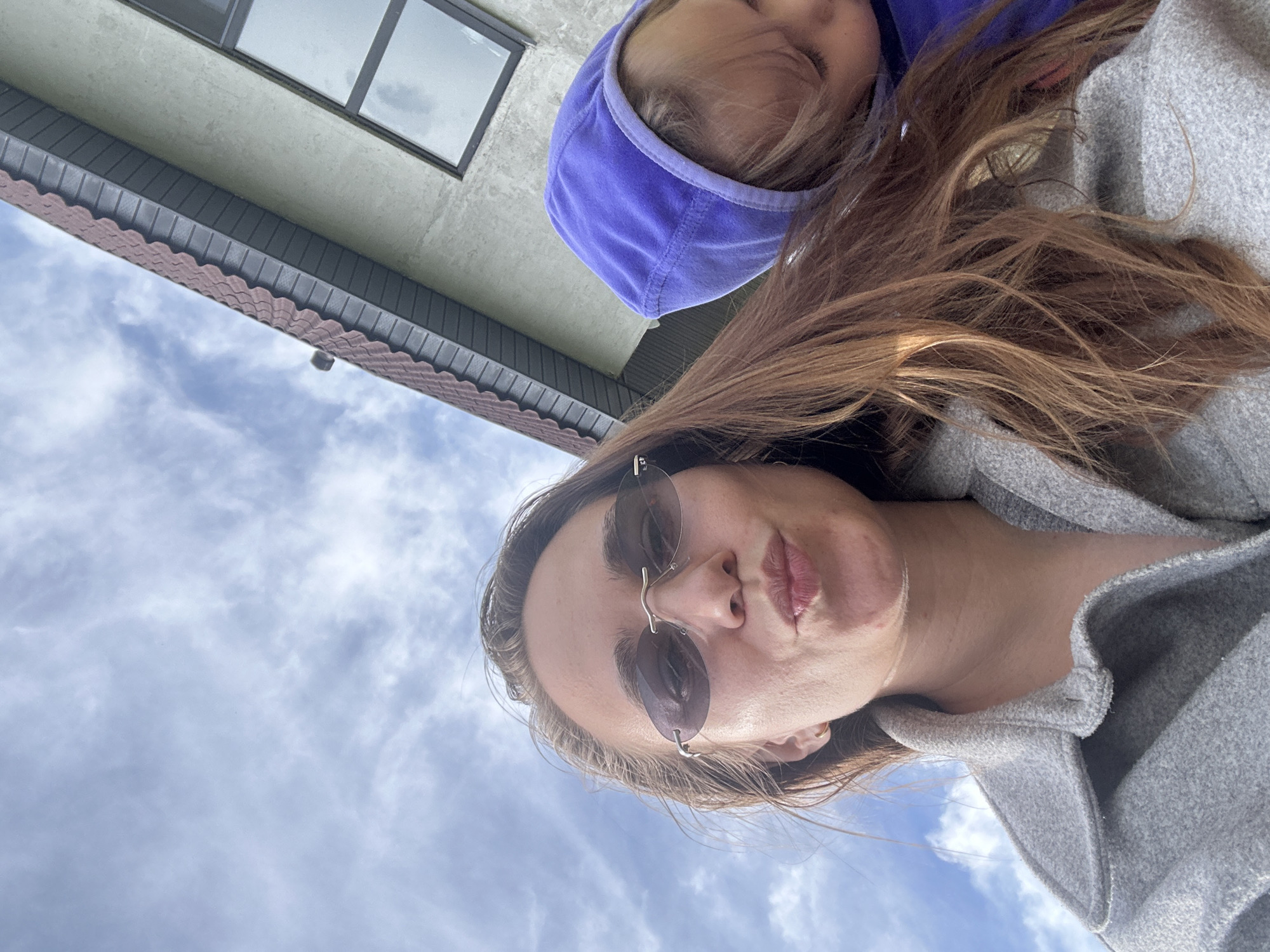 This low-angle, sideways-oriented image features an adult and a child against a sky and building backdrop in Kharnavy, Russia. In the foreground, an adult female with long brown hair wears rimless sunglasses and a grey, textured coat, looking towards the camera with a playful, pouting expression. A child, partially visible above and behind the adult, is wearing a bright purple hooded top, with only a portion of their face and head visible.
The background consists largely of a bright, cloudy blue sky. On the left side of the frame, a building is visible, featuring a light-colored wall (possibly stucco) with dark-framed windows. Below the windows, there's a dark eaves structure, followed by a distinctive reddish-brown, scalloped decorative trim, likely part of the roofline or siding. The overall impression is a daytime scene with overcast but bright weather. No text is visible in the image.