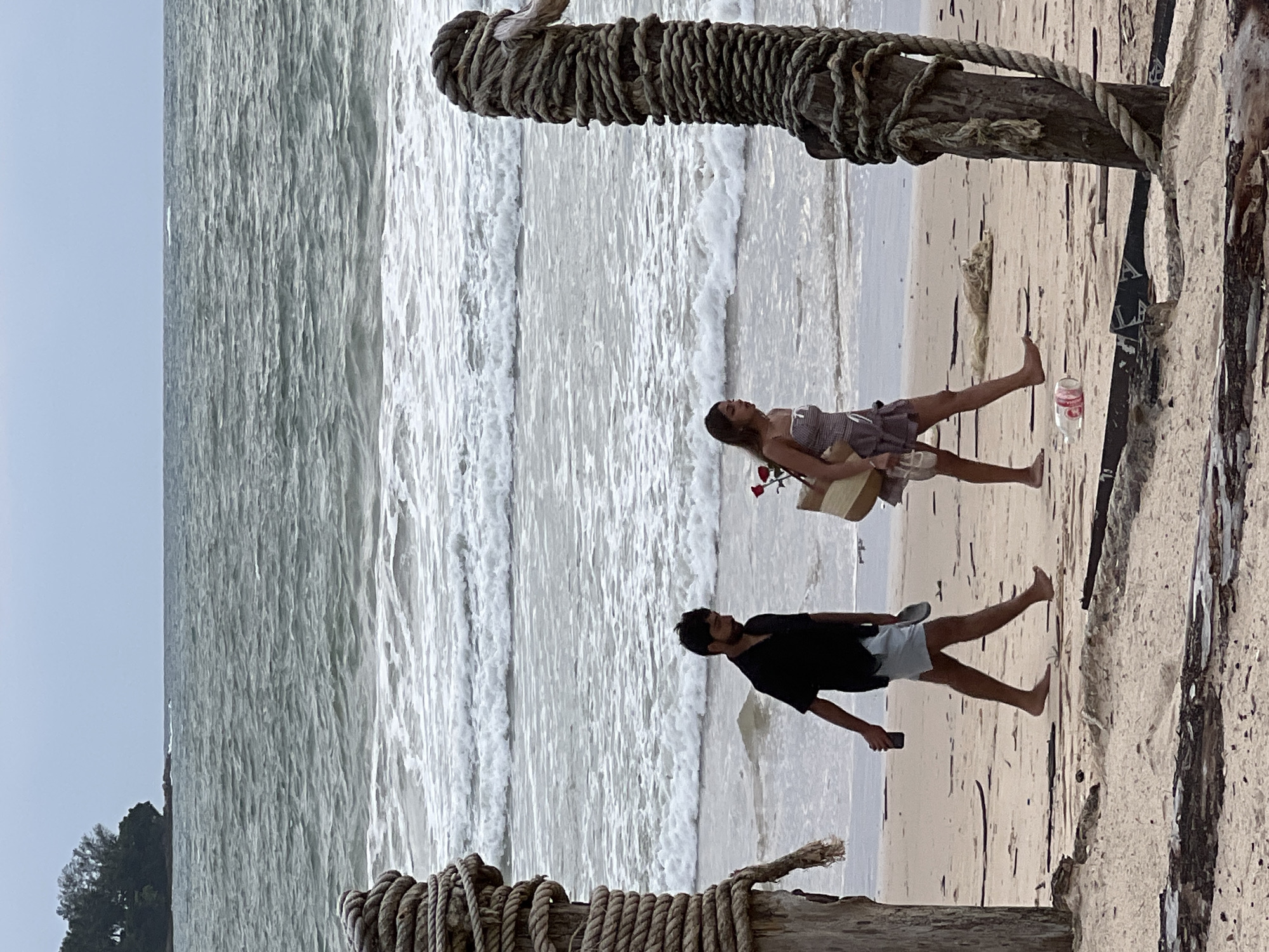 The image depicts a beach scene with two people on the sand. In the foreground, a man wearing a black t-shirt and light-colored shorts is walking, holding a dark object, possibly a phone, in his right hand. Behind him and slightly to his left, a woman in a patterned dress is also walking. She is carrying a light-colored bag and holding a red rose. The ocean with waves can be seen in the background, and a wooden structure wrapped in rope is prominently featured in the foreground and upper portion of the image. The sand appears wet near the water. The lighting suggests it could be daytime, with overcast or hazy conditions. The mood appears relaxed and perhaps romantic, given the presence of the rose. There is no visible text in the image.
