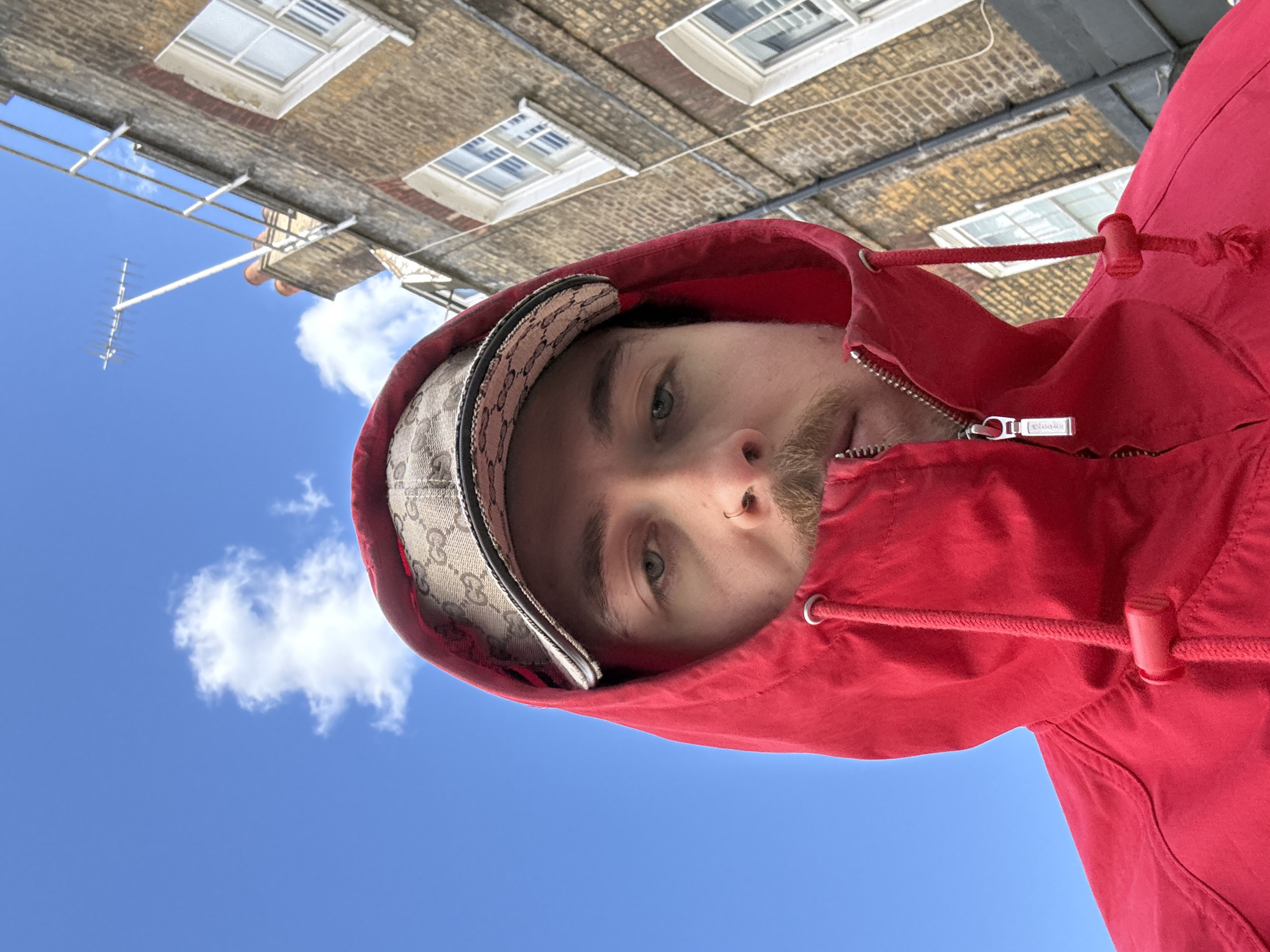 This media file presents a portrait of a young man, captured from a low angle and rotated 90 degrees counter-clockwise. The man's face, visible from the mid-forehead to just below the nose, looks downwards towards the camera. He has light-colored eyes, light facial stubble, and a small ring in his left nostril. He is wearing a bright red hooded jacket, the hood pulled up around his face, and a beige baseball cap with a distinctive dark brown monogram pattern, reminiscent of Gucci. His expression is neutral.
The background is dominated by a clear blue sky with a few scattered white clouds, indicating bright daylight and sunny weather. Above the clouds and the man's head, a multi-story brick building with several windows is visible, appearing upside down due to the image's orientation. The building features light-colored window frames and sills, and an old-style brick facade typical of an urban environment in London, United Kingdom. A television aerial or antenna is also visible in the sky.
Notable details include the silver zipper pull on the red jacket, which clearly displays the text "RALPH LAUREN". The drawstring of the hood is also visible. The image appears to be a casual selfie, utilizing an unusual upward perspective against the cityscape.