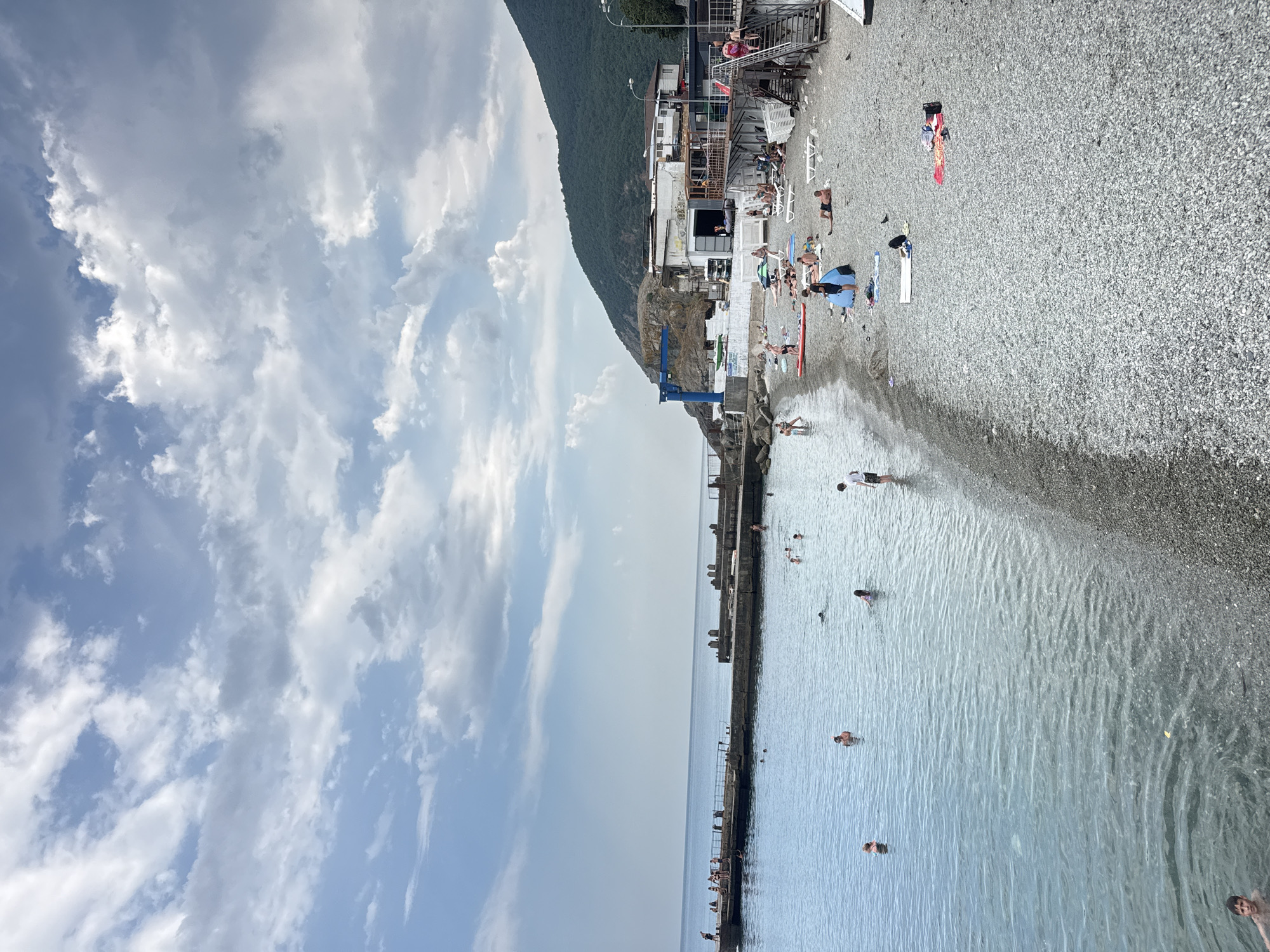 The image depicts a rocky beach with the Black Sea on the left and a steep, tree-covered mountain on the right. A concrete pier extends into the sea from the left side of the frame, with several people standing and sitting on it. The water is clear and calm, with numerous people swimming and wading. The beach itself is populated with sunbathers relaxing on towels and lounge chairs. Several buildings, including what appears to be a cafe or restaurant, are situated on the edge of the beach, at the base of the mountain. A large blue crane is visible behind some of these structures. The sky is partly cloudy with dramatic formations. The scene suggests a typical summer day at a popular beach resort in Partenit, Crimea, Ukraine.