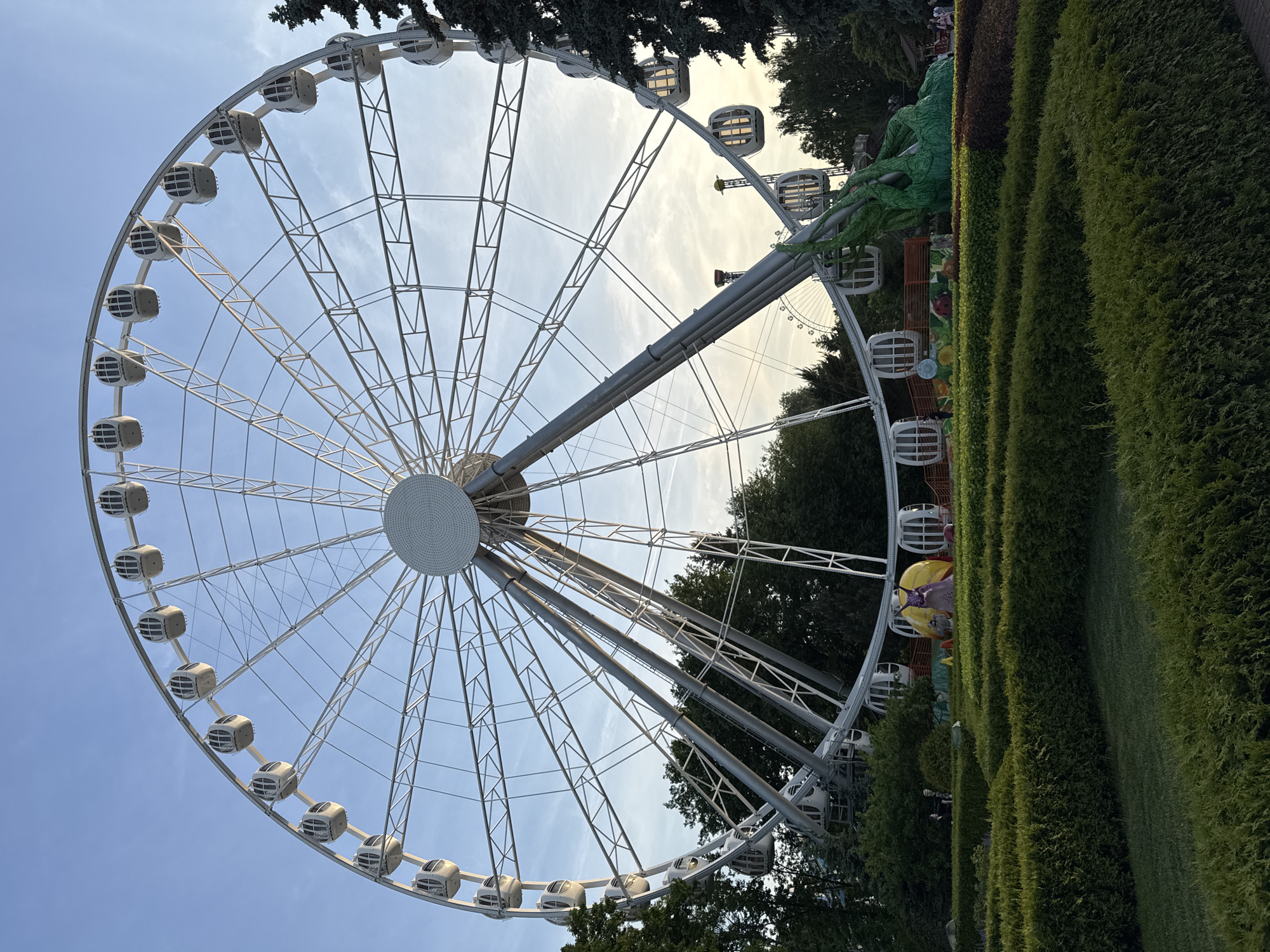 The image shows a large Ferris wheel at an amusement park. The Ferris wheel is white and has many cabins. In the foreground, there are manicured hedges and a labyrinth-like garden. Behind the hedges, there are some amusement park attractions, including a yellow structure and a large green dragon sculpture. In the background, there are trees and a sky with some clouds. There are a few people visible in the background, walking around the park. The overall scene suggests a pleasant day at an amusement park. This Ferris wheel is located in Saint Petersburg, Russia.
