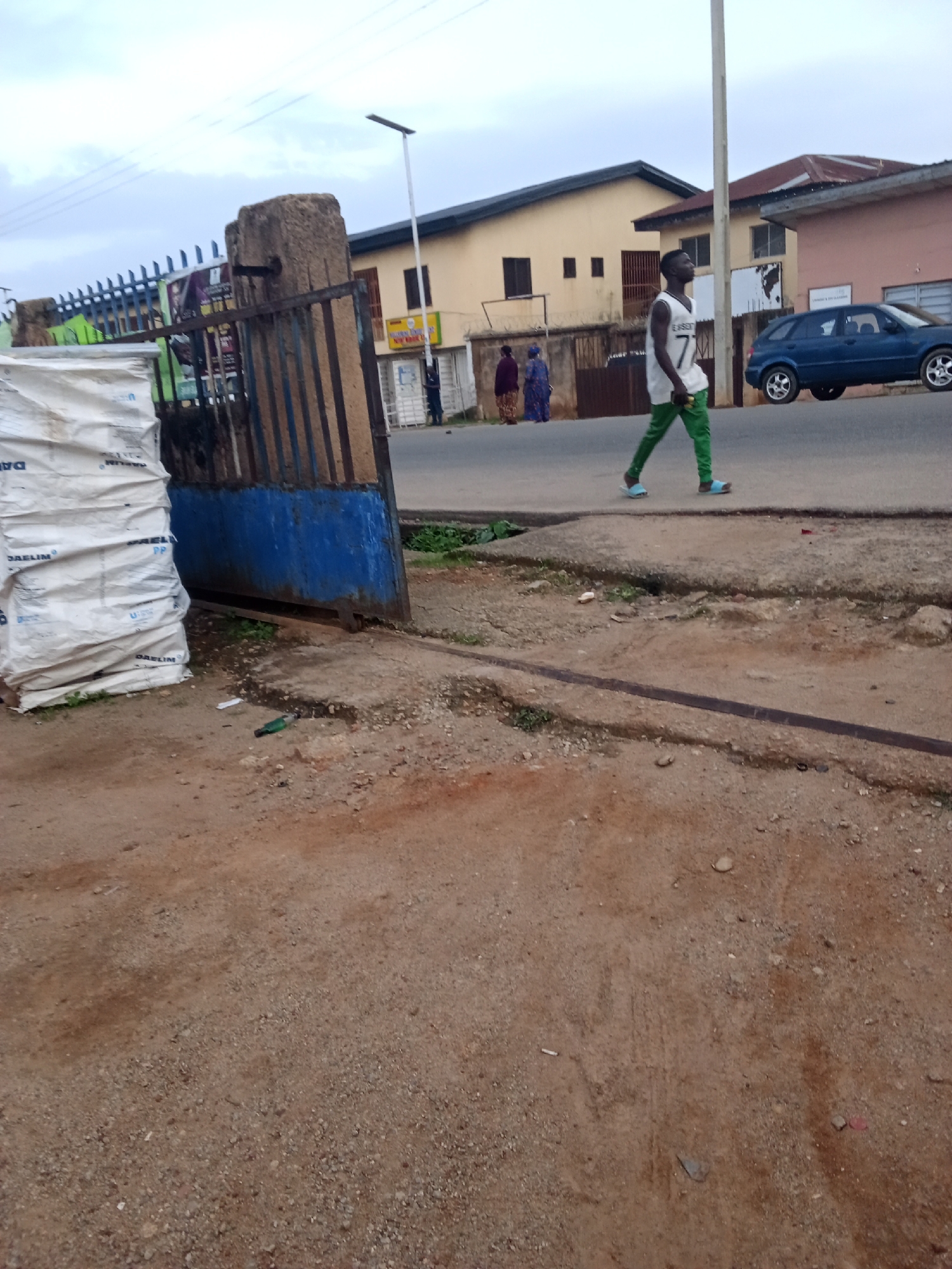 A full outdoor shot captures a street scene in Jos, Nigeria, with a pedestrian walkway in the foreground and a road to the right. A young man in a white tank top with the number "7" and green trousers walks on the walkway, facing the left of the frame. Behind him, two women are visible, walking away from the camera. A blue SUV drives on the road. The background features buildings with tan and pink facades, and a tall lamppost with a solar panel stands near the road. A large stack of white bags, possibly industrial materials, is positioned to the left of a rusty metal gate. The ground is predominantly dirt and gravel.