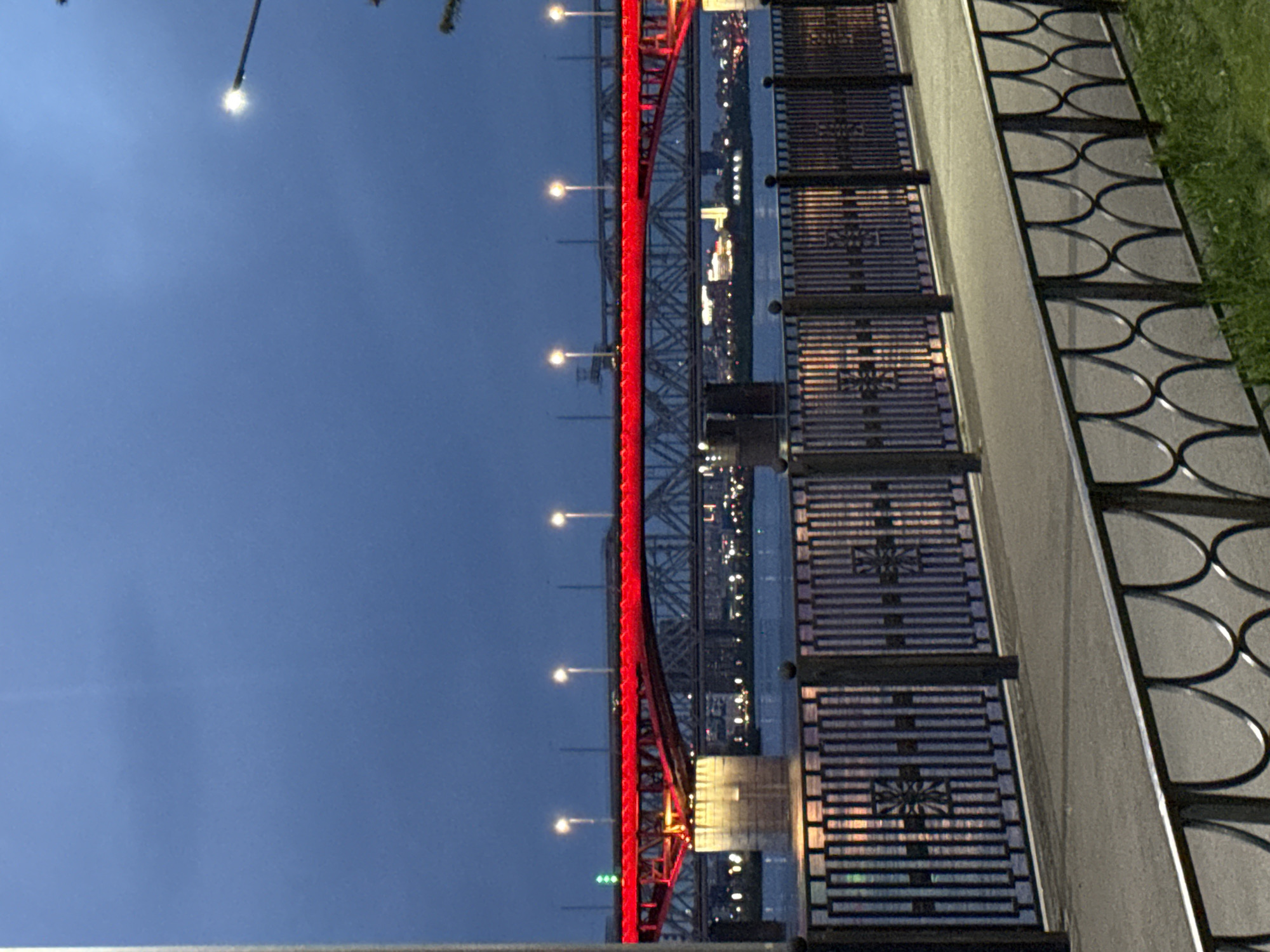 The image shows a night view of the Vinogradovsky Bridge in Krasnoyarsk, Russia, captured from a pedestrian walkway. The bridge is illuminated with red lights along its structure and features streetlights. The bridge spans a body of water, reflecting city lights. The pedestrian walkway has a decorative metal railing with circular designs. A section of grass is visible beside the walkway. The sky is a dark blue.