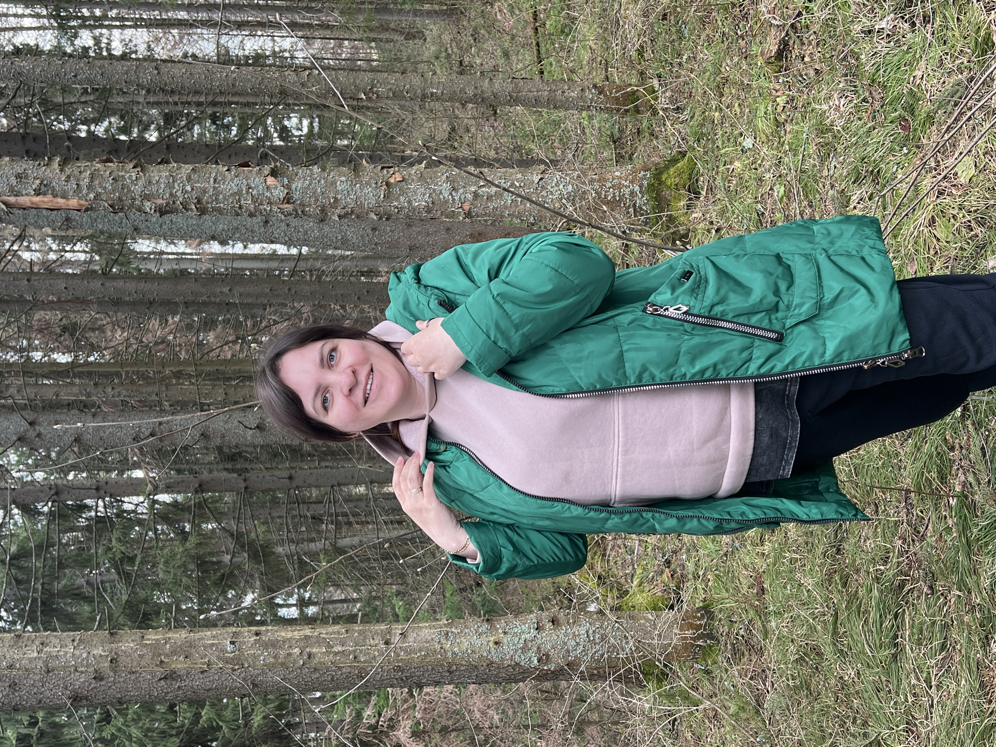 A woman is standing in a forest in Bol'shakino, Russia. She is wearing a green puffer jacket over a light pink hoodie and black pants. The forest is characterized by tall, thin trees and ground cover of dry grass and twigs.