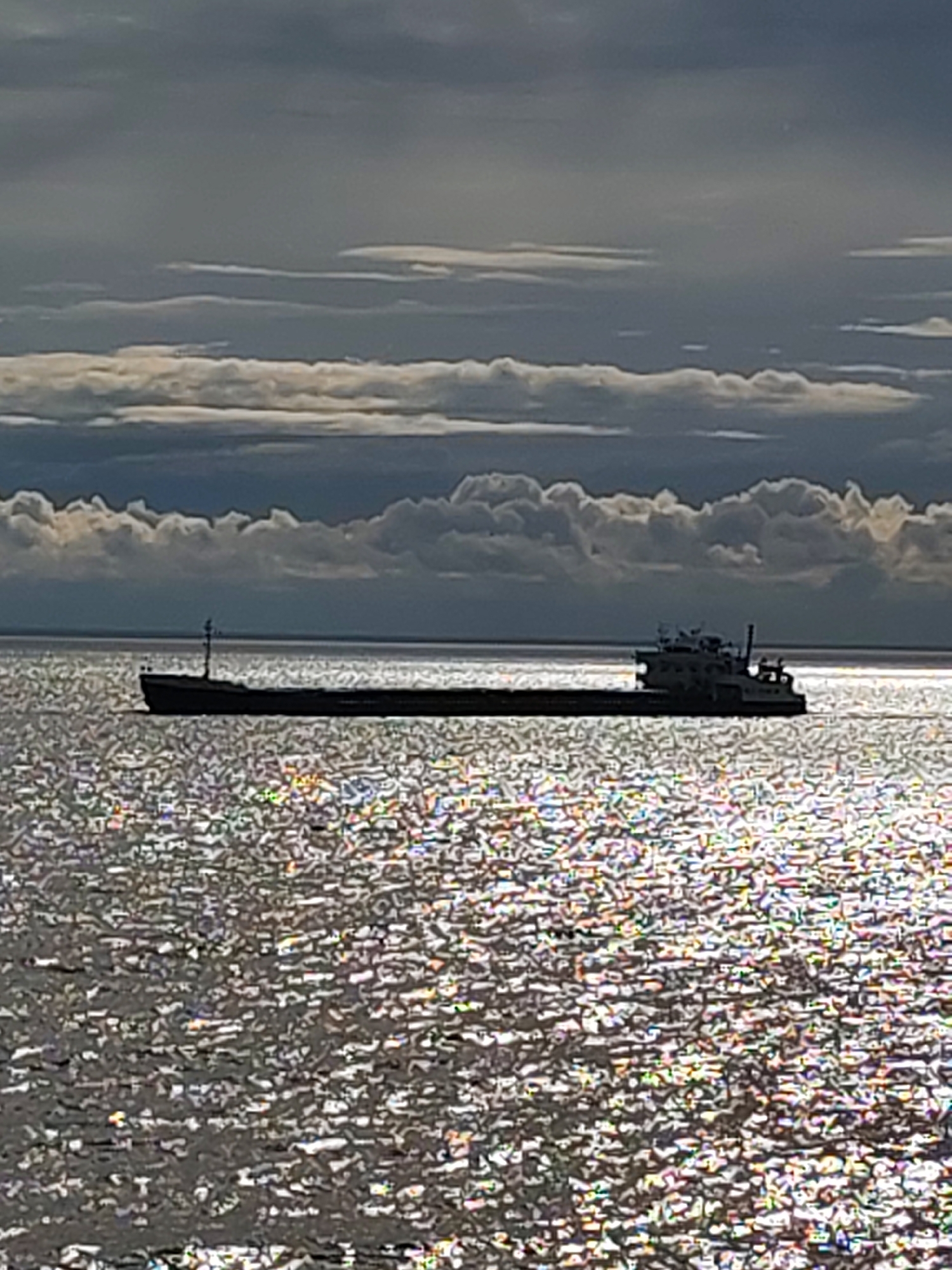 A medium-distance shot shows a long barge floating on a choppy body of water under a cloudy sky. The barge is black, with a taller structure at its rightmost end and a pole at its leftmost end. The water is a shiny, speckled gray color due to the sun's reflection. The sky is filled with thick, gray clouds in several layers, the layers toward the horizon whiter than those above. The scene is Belozersk, Russia.
