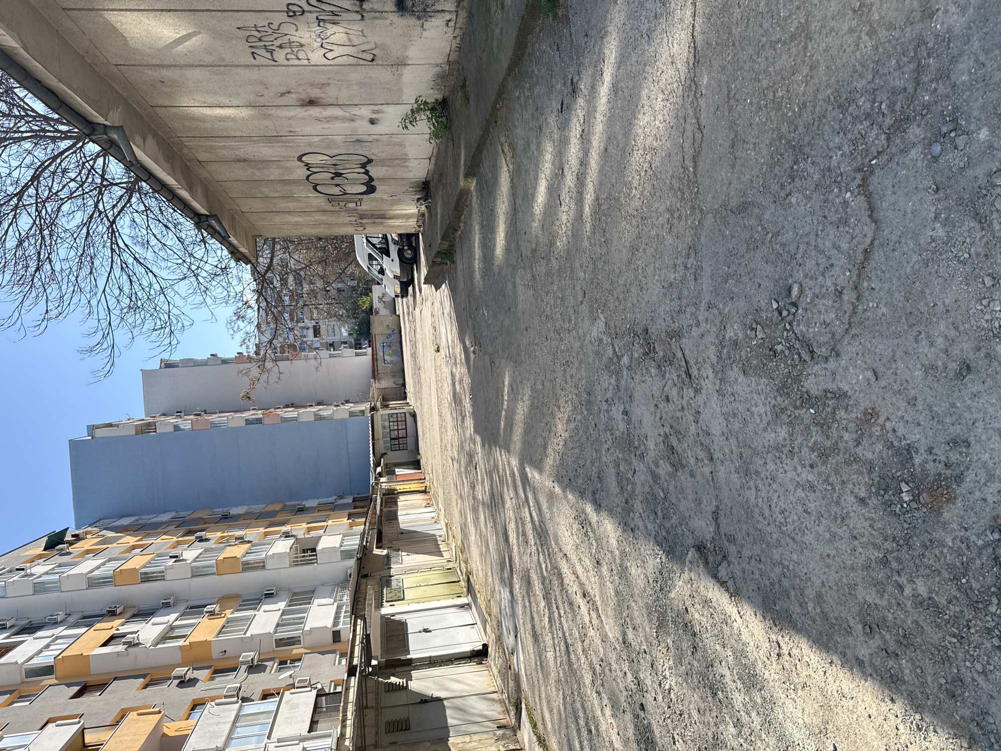 The image depicts an urban alleyway in Stara Zagora, Bulgaria. The alley is paved with cracked and worn concrete. A concrete overpass or bridge runs along the upper left of the frame, covered in graffiti. Along the left side are tall apartment buildings, one with a predominantly yellow and white facade, and another with a blue section. The sky is visible in the upper left corner and is clear blue. A bare tree branch is visible against the sky. A white van is parked further down the alley. The alley floor is marked by shadows, indicating sunlight.