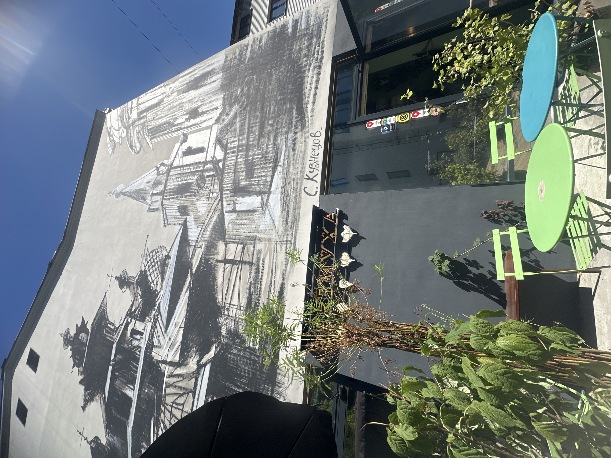 The image shows a street-level view of a building with a large mural on its facade, located in Moscow, Russia. The mural, rendered in grayscale, depicts an architectural scene with multiple towers and ornate structures, suggestive of historical Russian architecture. Below the mural, a dark-colored building facade features a glass entrance with several small circular stickers on the glass. In front of this facade, there is an outdoor seating area with two bright green metal tables and chairs. A large potted plant with green foliage and thin, brown stalks is positioned prominently in the foreground, partially obscuring the tables and chairs. To the left of the seating area, a large black umbrella is visible, casting a shadow. The sky is a clear, bright blue. The overall scene suggests a cafe or restaurant with outdoor seating situated in an urban environment.