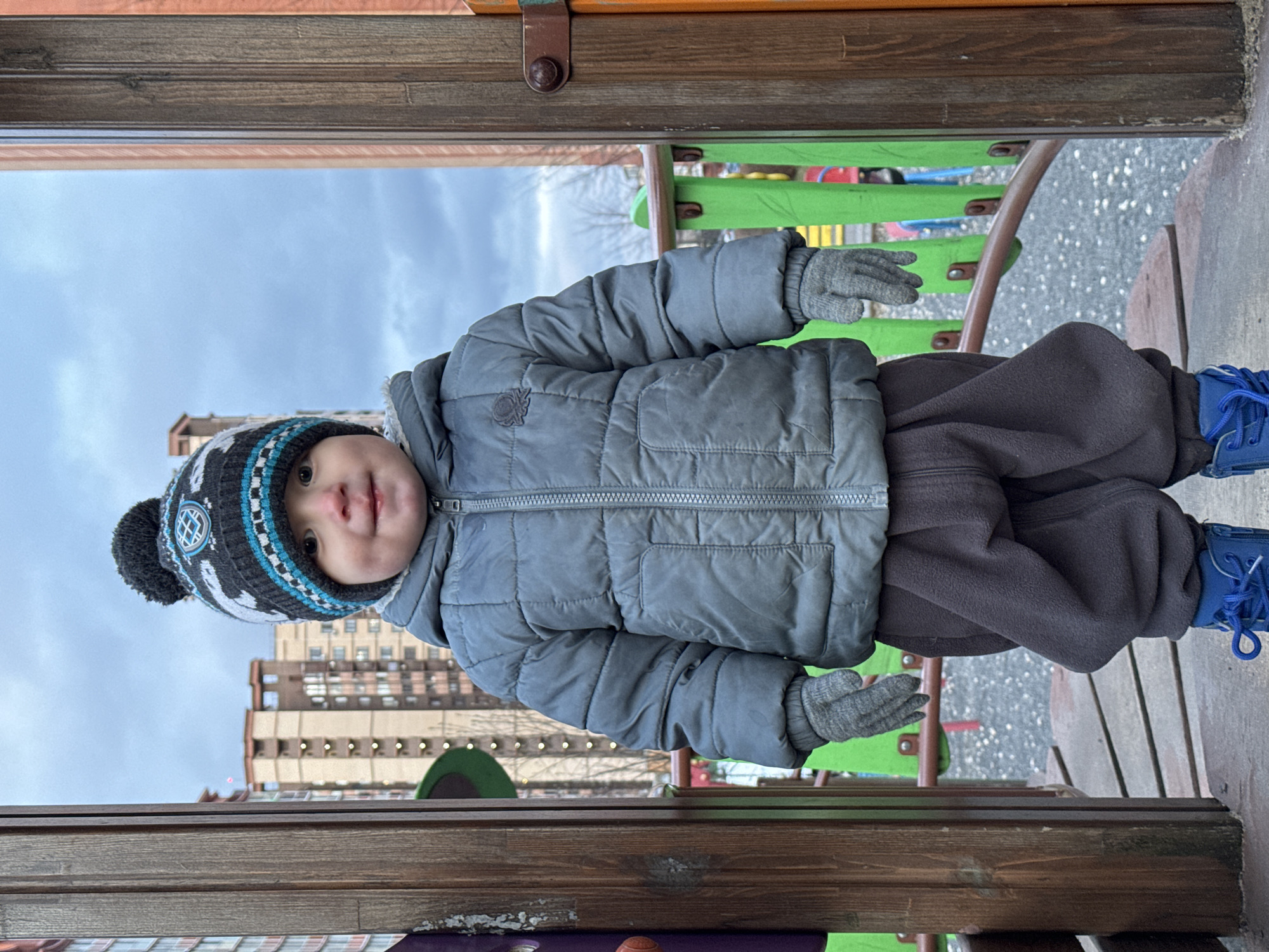 A child in winter clothing stands on a playground structure. The child wears a gray puffer jacket, dark pants, a patterned winter hat with a pom-pom, gray gloves, and blue shoes. The background shows buildings, possibly in the city of Odintsovo, Russia, based on architectural styles. The playground includes wooden and green metal components. The sky is overcast. The child appears to be looking directly at the camera.
