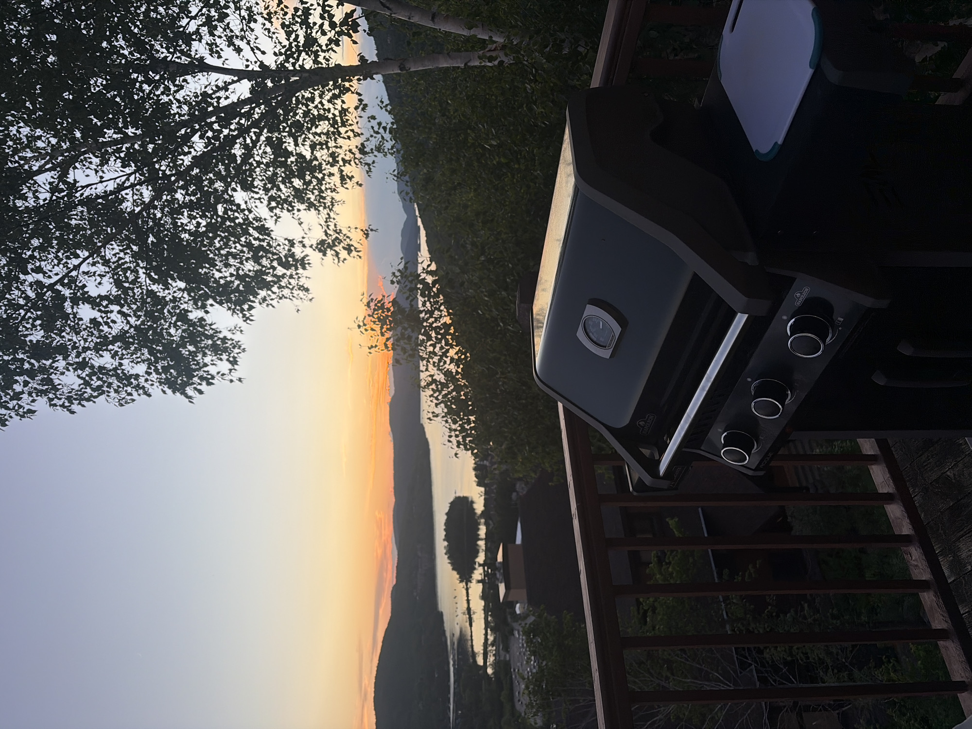 This image depicts a gas grill on a wooden deck overlooking a body of water and rolling hills at sunset. The sky displays a gradient of soft oranges and blues, with the last rays of sunlight illuminating the landscape. The grill, a Napoleon brand, is positioned in the foreground, its lid closed, revealing a thermometer on the front. To the right of the grill, a white cutting board with a green trim rests on the deck. The deck itself is constructed of dark wood and is bordered by a wooden railing. Beyond the deck, lush green trees and foliage dominate the middle ground. In the distance, a large body of water is visible, with a small island in its center. Hills rise beyond the water, their contours softened by the twilight. The scene is likely located in or near Mont-Tremblant, Quebec, Canada, a region known for its natural beauty and outdoor recreational activities. No people are visible in the image.