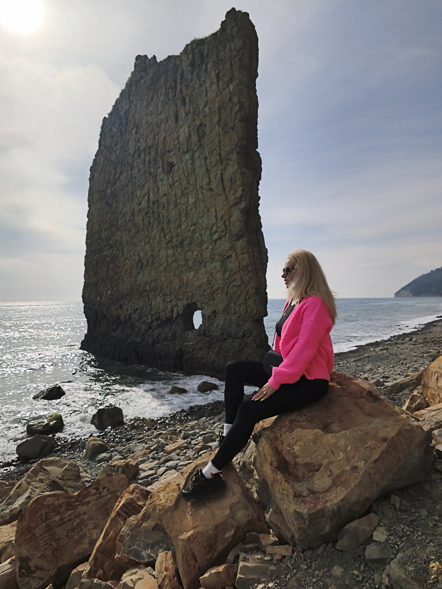 A woman with long blonde hair, wearing a bright pink cardigan, black leggings, and black sneakers with white socks, sits on a large brown rock on a rocky beach. She faces right, looking out towards the sea. Behind her is a tall, imposing rock formation with a distinct hole near its base, partially submerged in the water. Waves are visible around the base of the rock formation and along the shoreline. The beach is covered in pebbles and rocks of various sizes. In the distance, a stretch of coastline with trees is visible. The sky is overcast with the sun partially obscured. The location is Praskoveyevka, Russia, known for its unique rock formations along the coast.