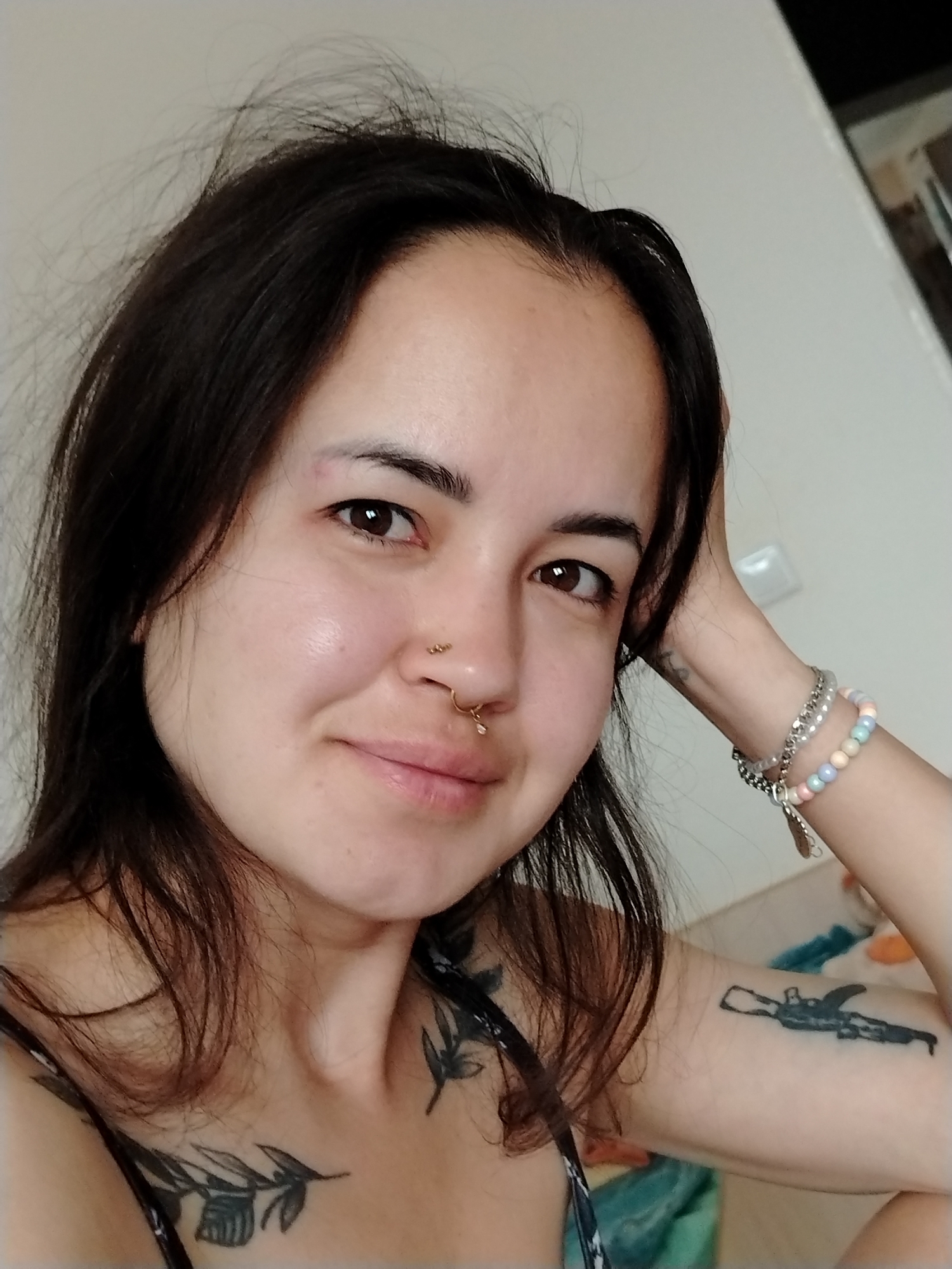 A woman is posing for a selfie. She has dark hair, brown eyes, and multiple nose piercings. Her left arm is raised with her hand touching the back of her head. Bracelets are visible on her left wrist. Tattoos are present on her left arm and chest area. The background shows a plain, light-colored wall and a glimpse of what appears to be a room interior. There is no indication of the location being in the city of Noviy, Russia.