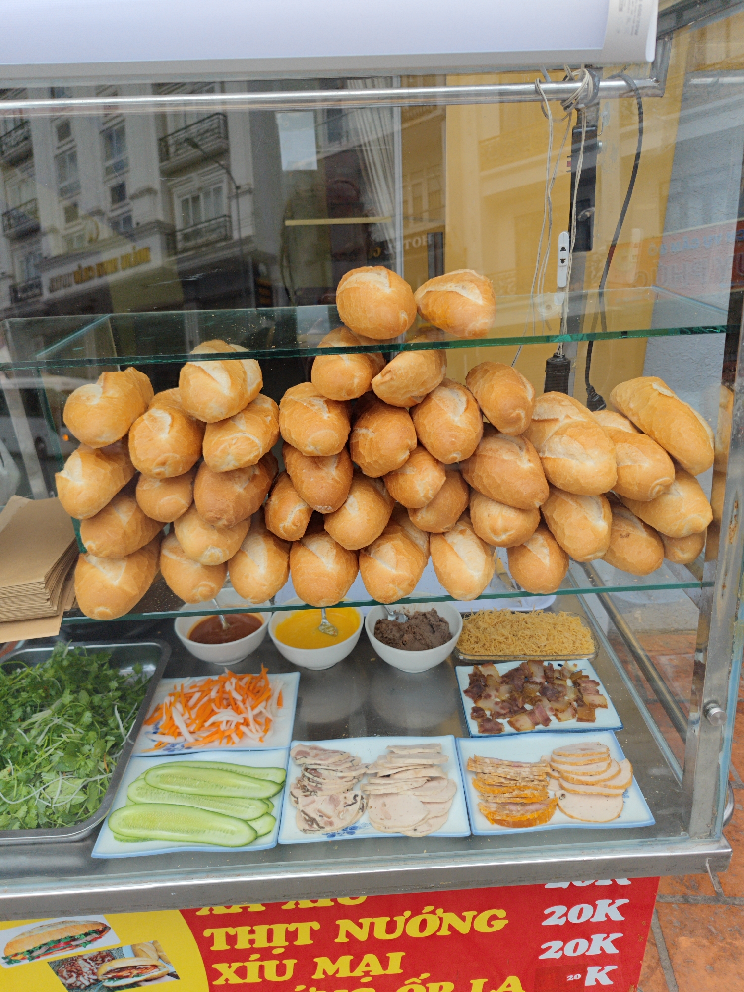 This vibrant close-up depicts a bustling Vietnamese street food stall, likely for Banh Mi sandwiches, set up in Da Lat. The scene is dominated by a glass display case filled with fresh ingredients and numerous golden-crusted baguette rolls.
On the top shelf of the display, dozens of small, roundish Banh Mi rolls are stacked high, ready for preparation. The middle section showcases an array of sandwich fillings: a metal tray overflowing with fresh green herbs (likely cilantro and mint), a plate of shredded pickled carrots and daikon, and another plate of sliced cucumbers. Adjacent to these are several plates holding different types of sliced cold cuts, including various pork rolls and what appears to be grilled or braised pork pieces. Small bowls contain sauces, one dark brown and another bright yellow (possibly mayonnaise or a butter spread), and a bowl of shredded pork floss.
No people are visible, but the meticulously arranged setup suggests an active vendor preparing for customers. The background, reflected in the glass, shows a multi-story building with balconies, indicating an urban setting. Bright, natural light suggests it is daytime. Clutter from electrical wires is visible attached to the display case's frame.
At the bottom, a red banner with yellow and white Vietnamese text and prices is prominently displayed. Visible text includes "THỊT NƯỚNG" (Grilled meat) and "XÍU MẠI" (Pork meatballs). Multiple instances of "20K" (20,000 Vietnamese Dong) denote the prices for various items. A small image of a finished Banh Mi sandwich is also shown on the banner, illustrating the final product. The overall impression is one of fresh, ready-to-assemble street food.