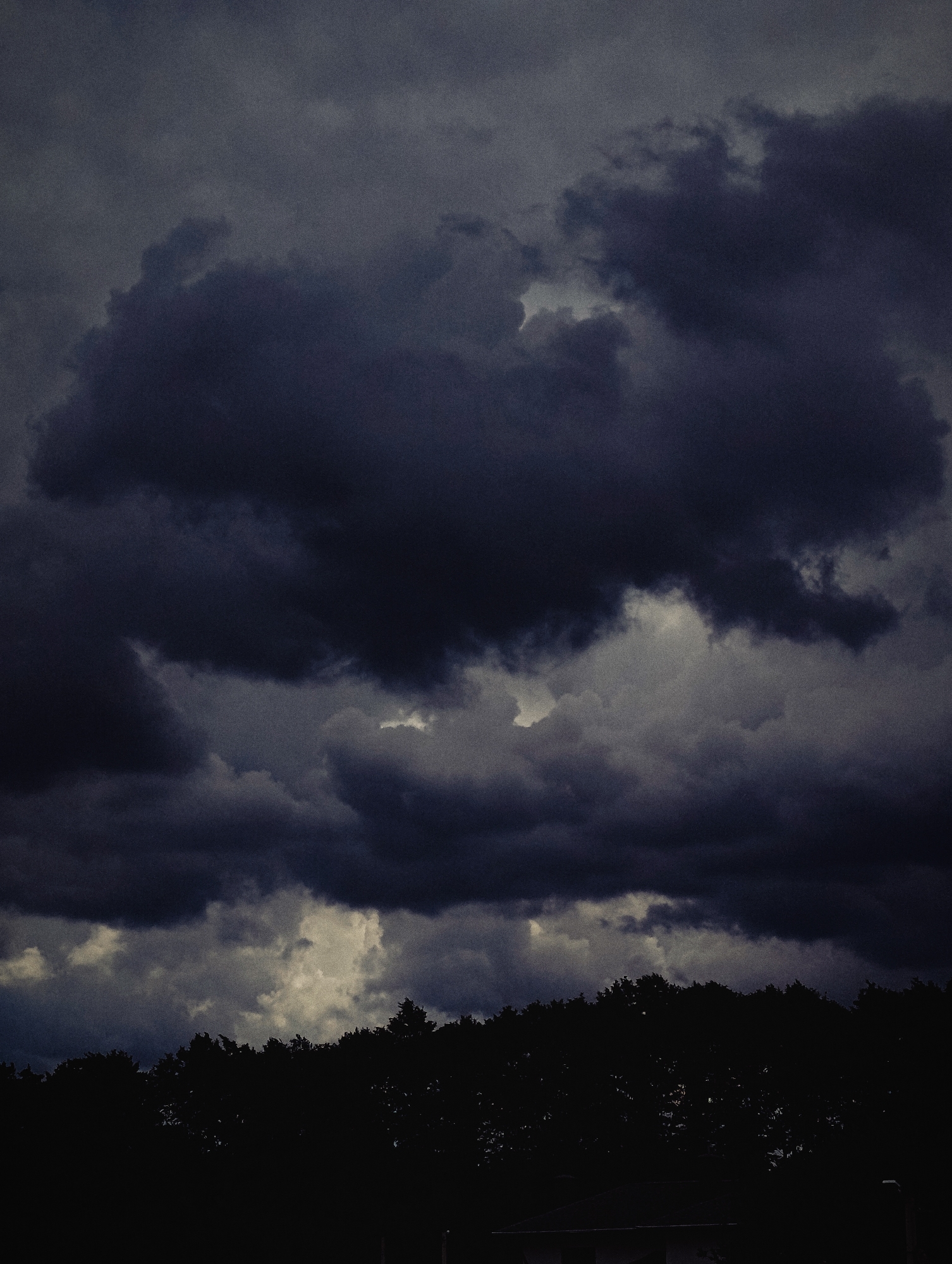 The image depicts a dark, overcast sky with heavy, turbulent clouds that dominate the upper portion. The clouds are a mixture of deep grays and blacks, with some lighter patches suggesting diffused light breaking through. Below the clouds, a dense line of trees forms a dark silhouette against the sky, obscuring the ground and any structures that might be present. The overall mood is somber and foreboding, indicative of impending bad weather. The image does not contain any specific landmarks or identifiable features that would pinpoint a location in Minsk, Belarus. There are no people, animals, or discernible objects in the scene. The environment is natural, characterized by clouds and a tree line.