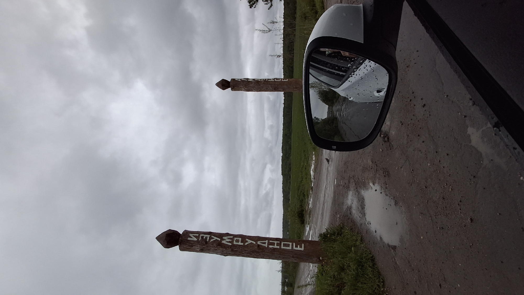A slightly high-angle, close-up outdoor shot depicts a gray car's side mirror capturing a view of a dirt road flanked by green vegetation and a forest edge under a cloudy sky. A weathered wooden signpost with a pointed top, inscribed with "NEMPAVAHOE" in white block letters, is visible both directly and in the mirror's reflection, indicating a location in Nempa, Russia. The road surface is wet, with puddles reflecting the overcast sky. The car's side panel is partially visible in the right edge of the frame.