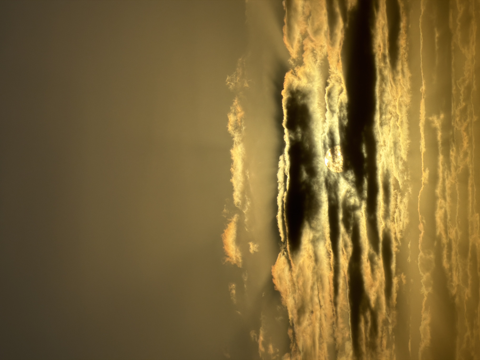 The image depicts a sky at sunrise or sunset, likely with the sun obscured by clouds. The sky transitions from a golden hue to a darker gray on the left. The clouds are vertically oriented, with dark, shadowy gaps between them, giving the impression of columns or streaks. The sun is partially visible through a break in the clouds. The exact location is not identifiable without additional context.