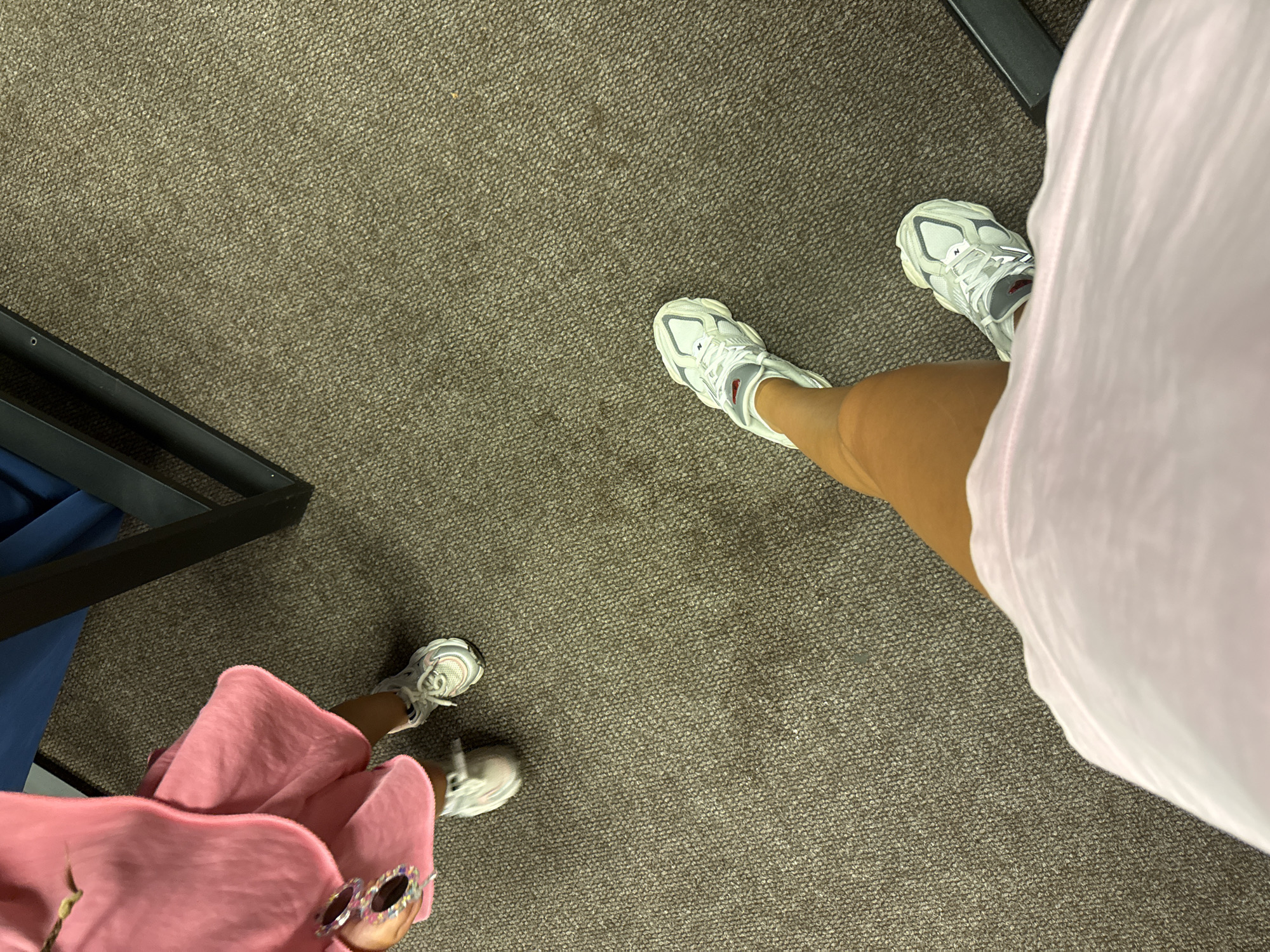 The image is a high-angle shot taken indoors, likely in a home or a fitting room. The flooring is a textured, neutral-colored carpet. In the upper left quadrant, a child is visible, wearing a pink outfit and white sneakers. The child is holding a pair of sparkly sunglasses. To the right of the child, a portion of a dark metal frame, possibly furniture or shelving, is visible. The lower half of the image focuses on the legs and feet of an adult, also wearing white sneakers. The adult is wearing a light pink garment, possibly a dress or a large t-shirt, that drapes over their legs. The scene appears to be casual and mundane, with no discernible elements suggesting a specific location within Chișinău, Moldova.