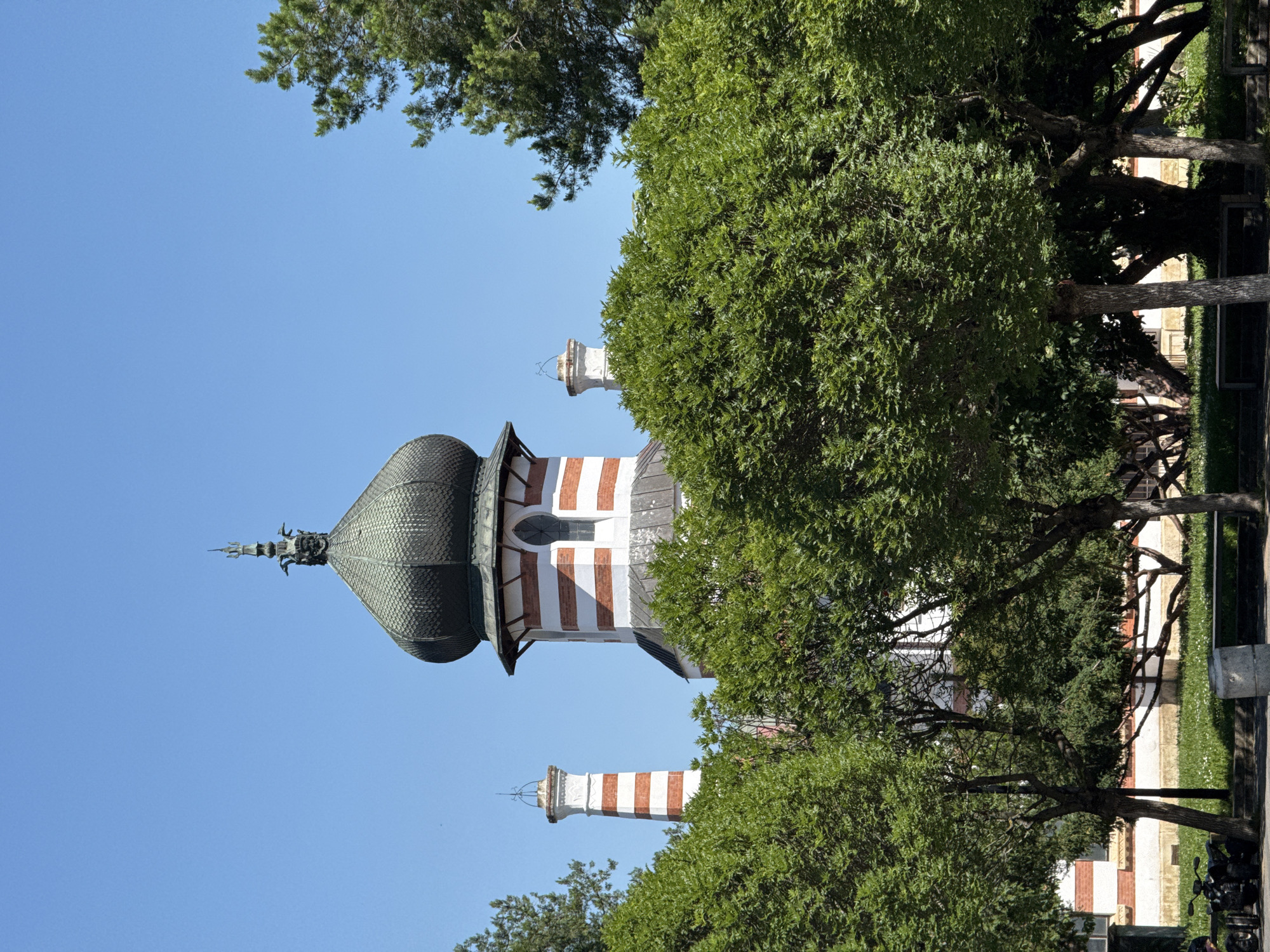 The image shows the Saint George the Victorious Mausoleum in Pleven, Bulgaria, from a low angle through surrounding trees. The mausoleum's dome is dark green, topped with a decorative metal spire. Below the dome, the structure is striped horizontally with white and reddish-brown bands. Parts of the mausoleum's towers are visible, also striped in the same colors. The sky is a clear blue, partially obscured by the trees surrounding the mausoleum. The trees are leafy, and their branches frame the building. Some sections of the park area surrounding the monument can be seen on the right side. The image is rotated 90 degrees counter-clockwise.